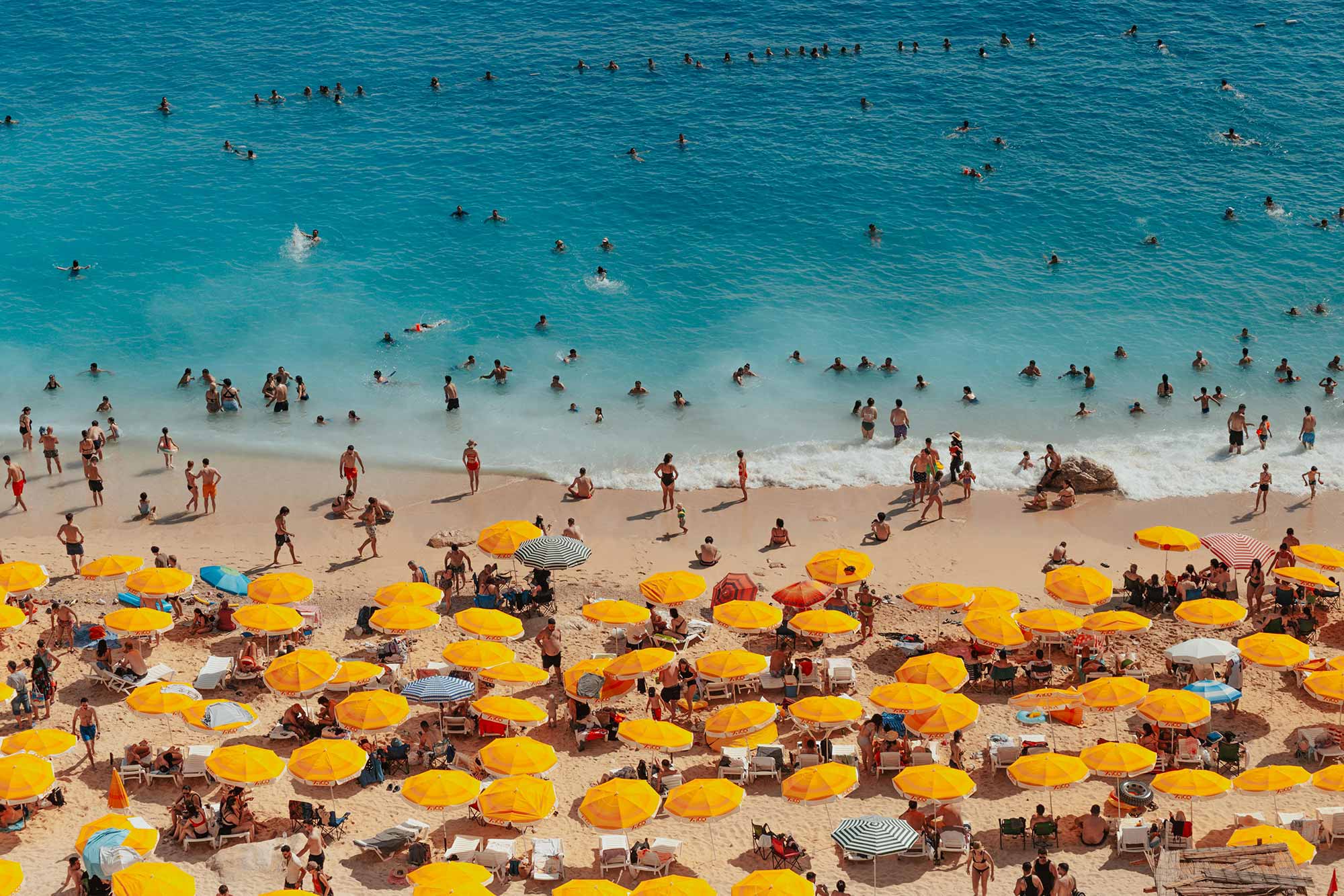 Luftaufnahme eines überfüllten Strandes mit gelben Sonnenschirmen und vielen Badegästen im türkisblauen Meer