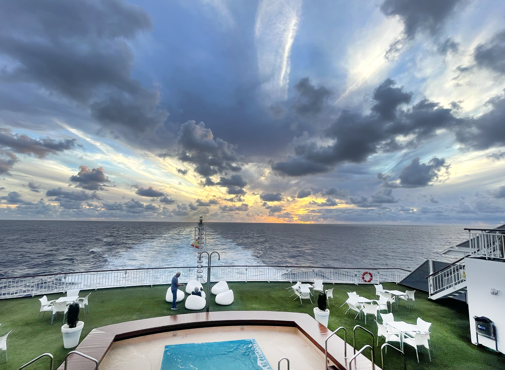 Achterdeck der Fähre mit Pool und weißen Loungemöbeln bei dramatischem Sonnenuntergang über dem Atlantik