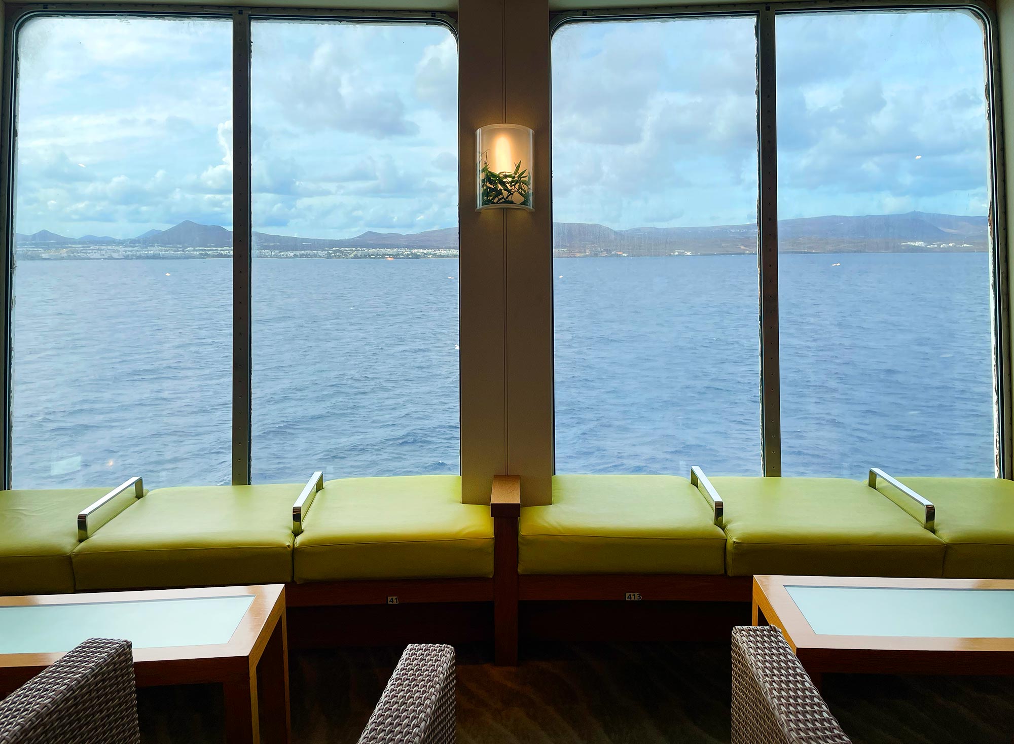 Loungebereich der Fähre mit gelbgrünen Sitzbänken, durch die großen Panoramafenster ist die Küste Lanzarotes zu sehen