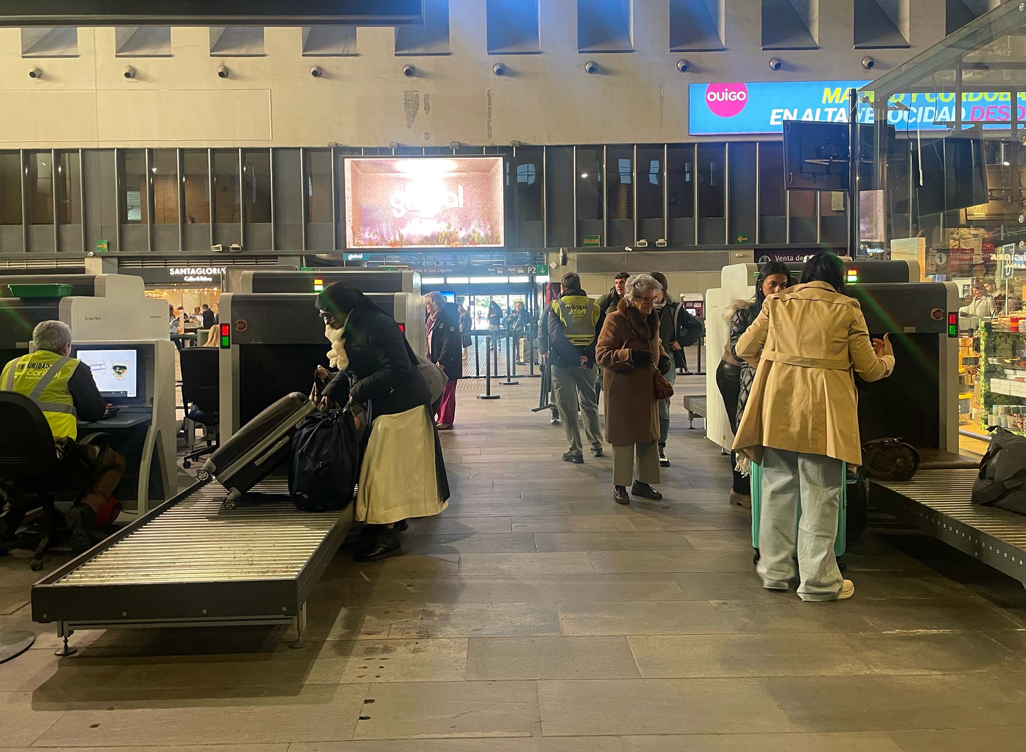 Sicherheitskontrolle im Bahnhof Sevilla mit Gepäckscanner und Reisenden, im Hintergrund Werbung für Hochgeschwindigkeitszüge