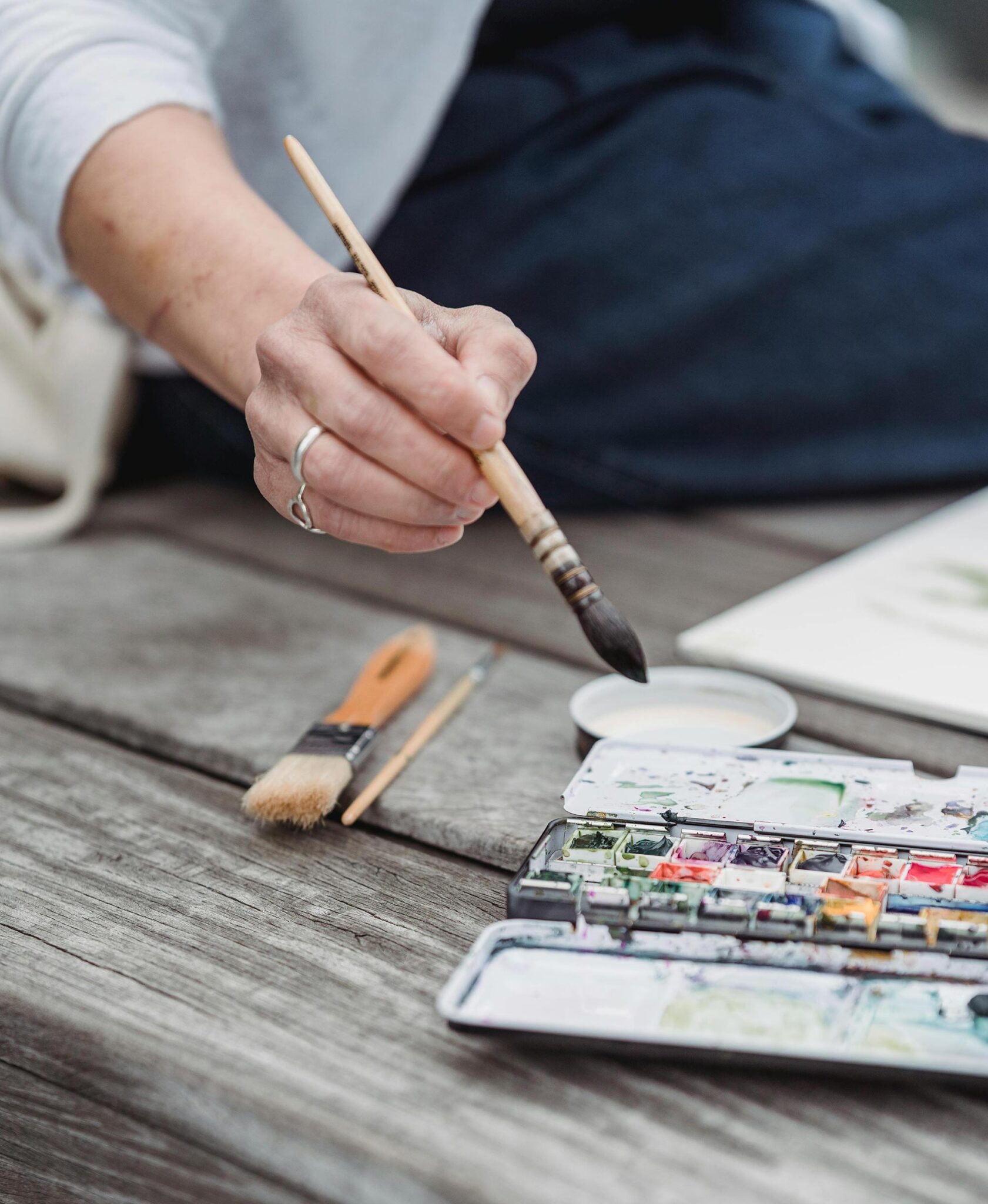 Hand mit Aquarellpinsel über einem Farbkasten auf verwittertem Holztisch