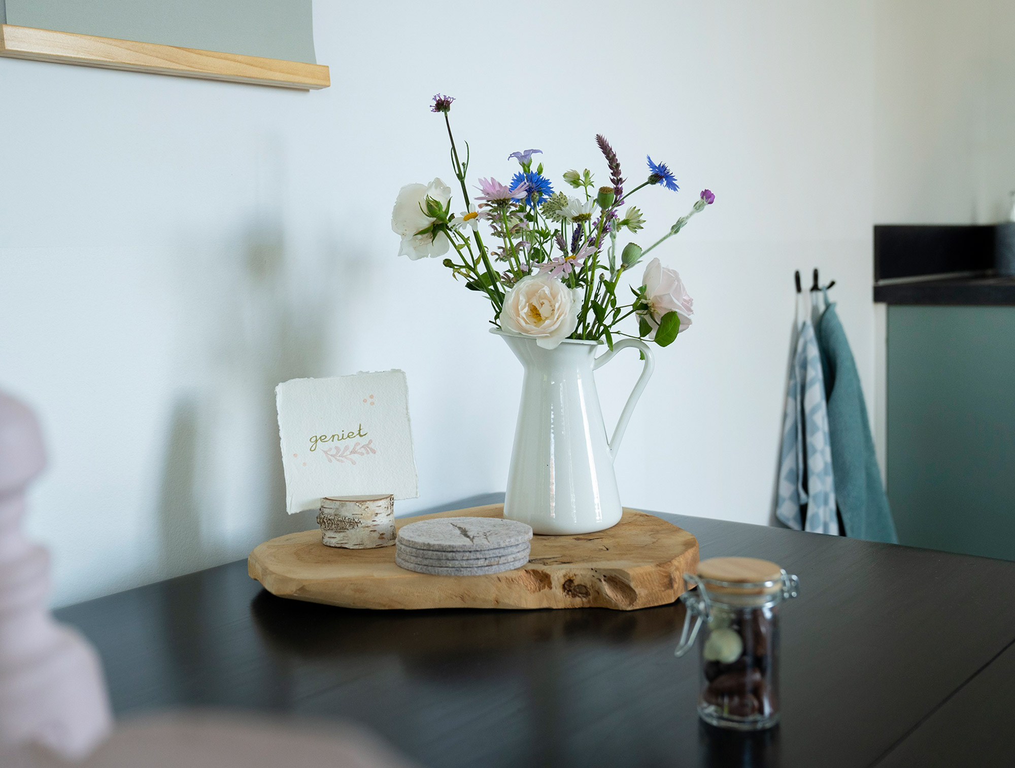 Tischdekoration mit weißer Keramikkanne, Wildblumenstrauß, Holzbrett und Karte mit niederländischem Schriftzug „geniet" – Zimmerdetail im B&B De Brenschutte, Niederlande