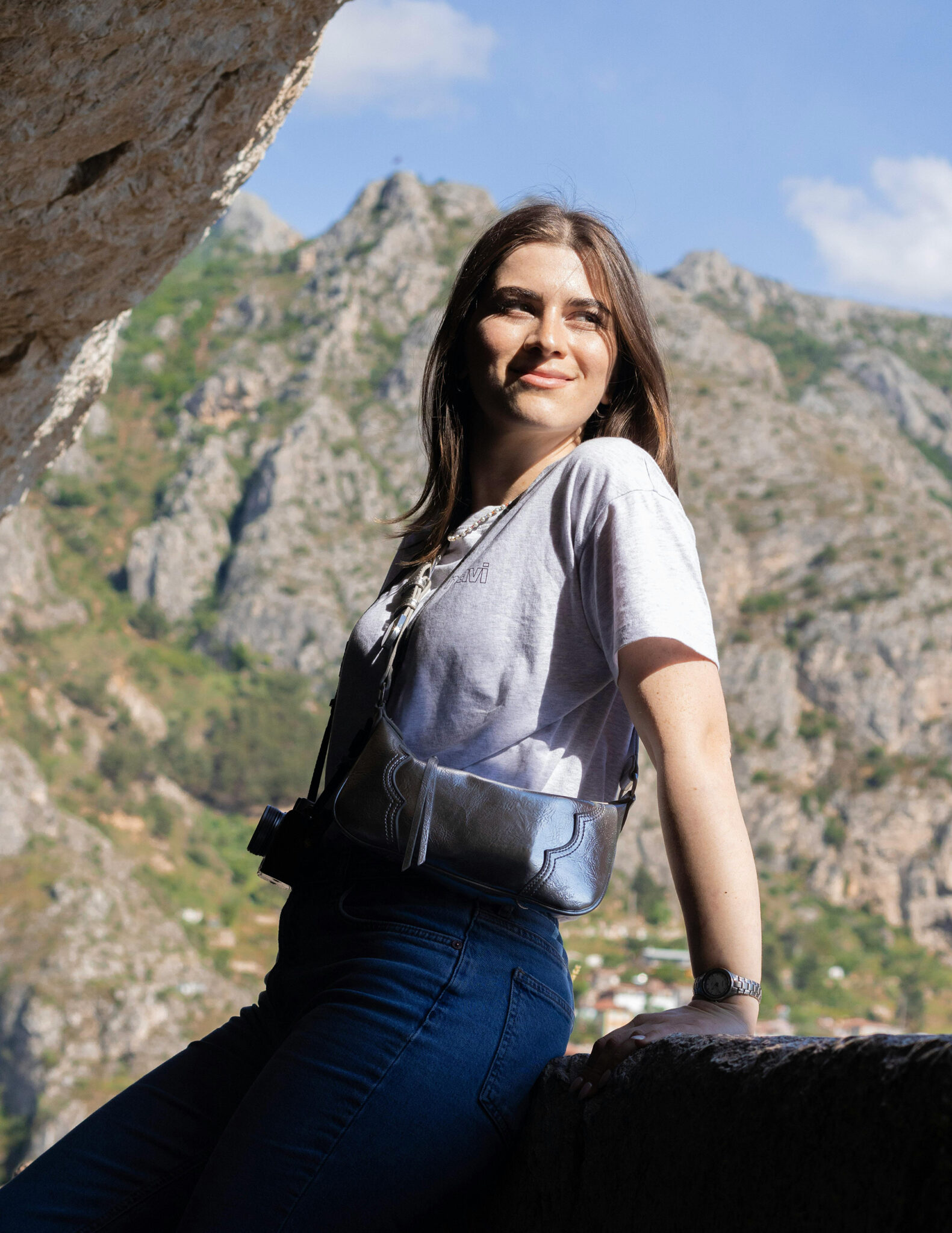 Lächelnde Frau mit Leder-Bauchtasche lehnt an einer Steinmauer vor imposantem Bergpanorama