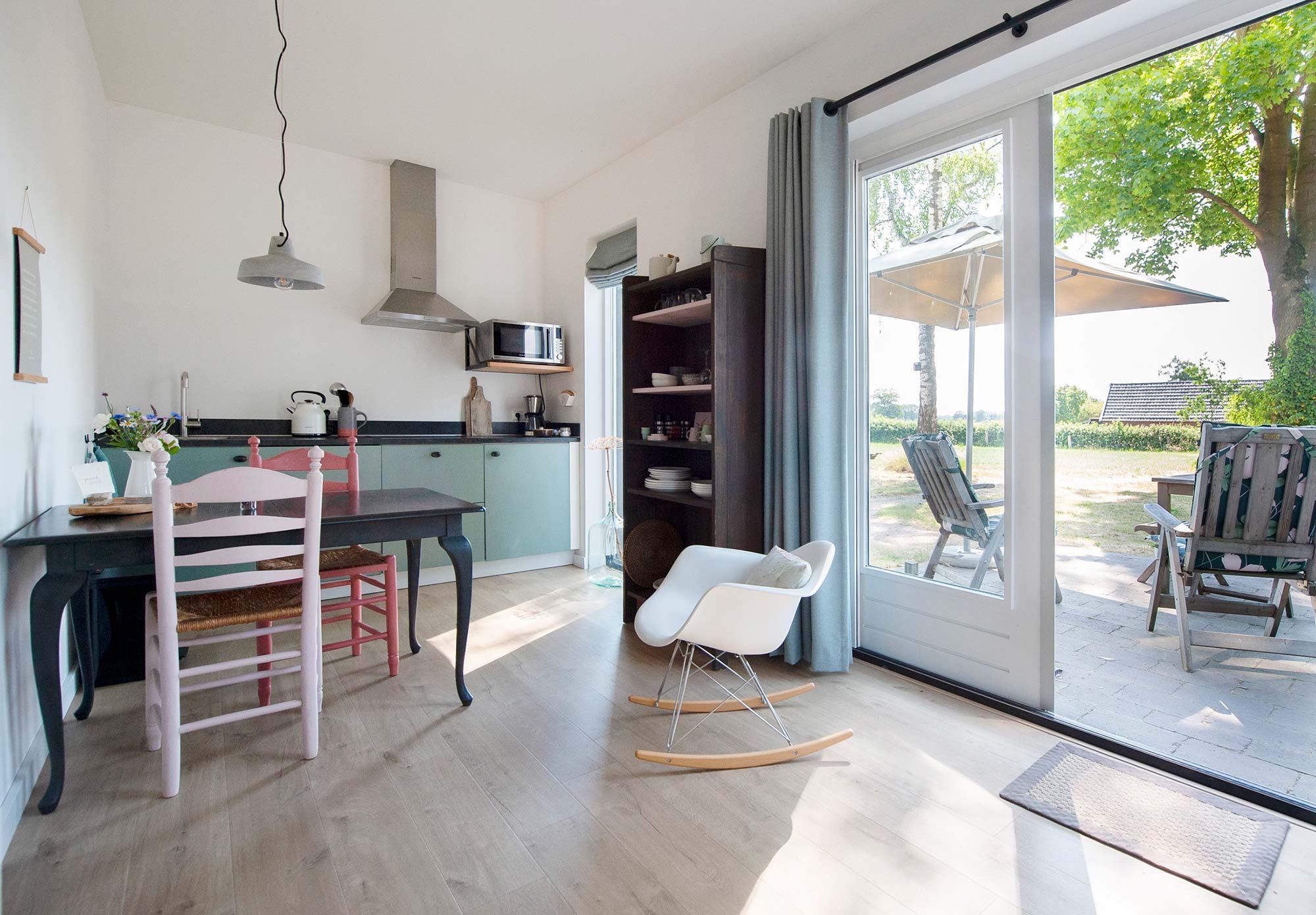 Helles Wohnzimmer mit Küche, rosa Stühlen, weißem Schaukelstuhl und großer Glasschiebetür zur Terrasse mit Blick ins Grüne – B&B De Brenschutte, Niederlande