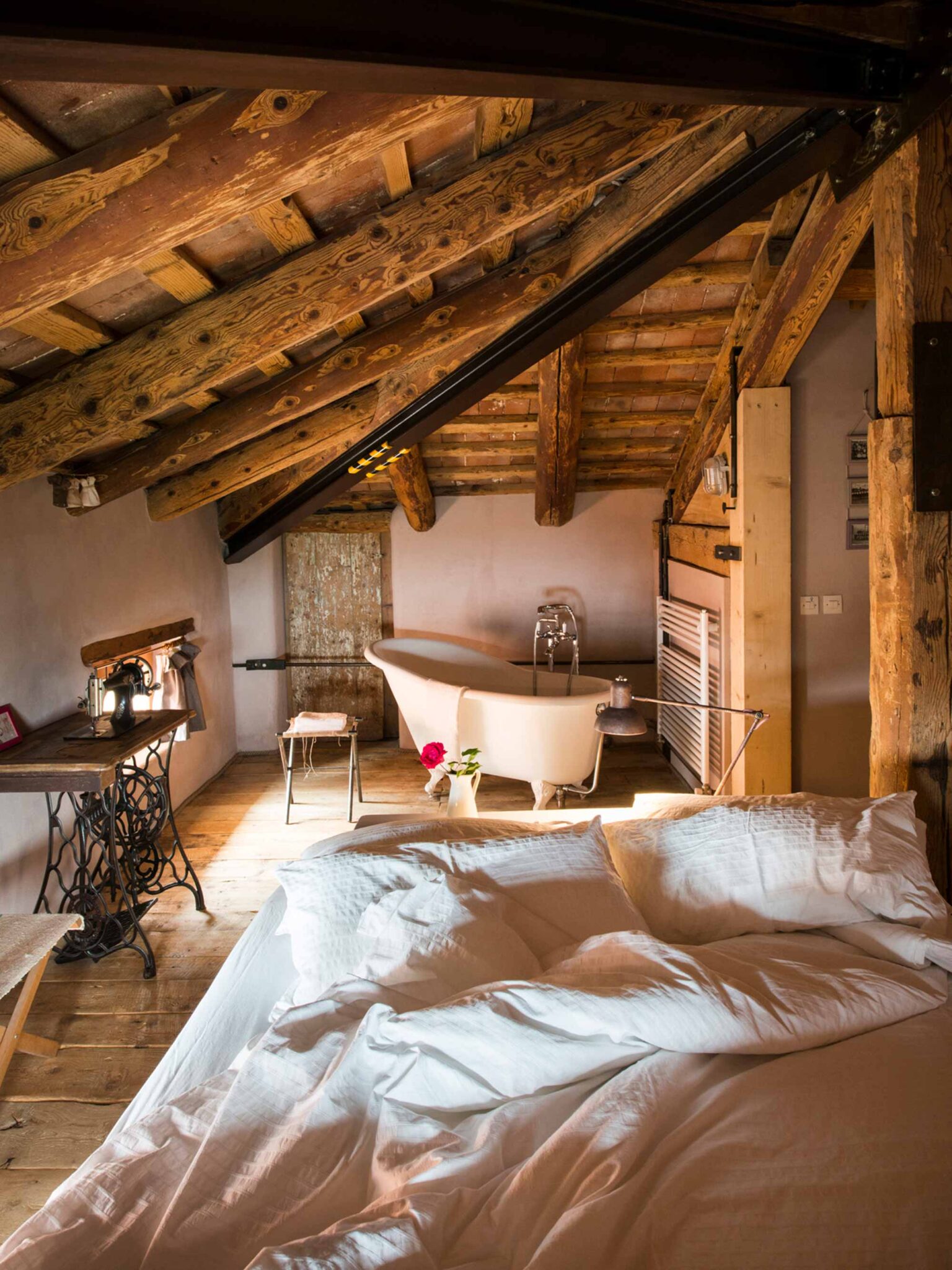 Rustikales Dachzimmer mit freiliegenden Holzbalken, freistehender Vintage-Badewanne und gemütlichem Bett mit weißer Bettwäsche