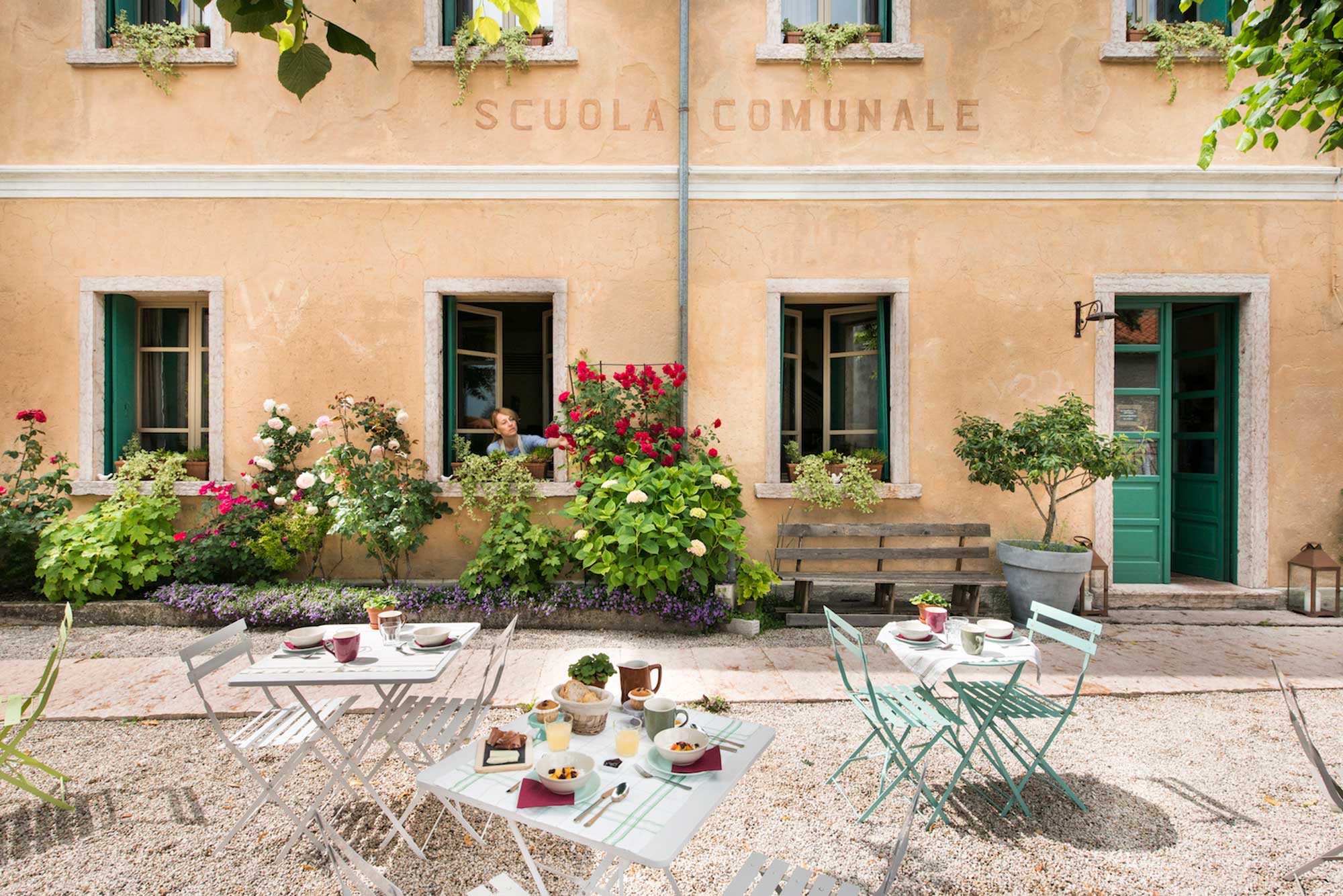 Historische Fassade des ehemaligen Schulgebäudes „Scuola Comunale“ mit grünen Fensterläden, blühenden Rosen und gedecktem Frühstückstisch im Garten