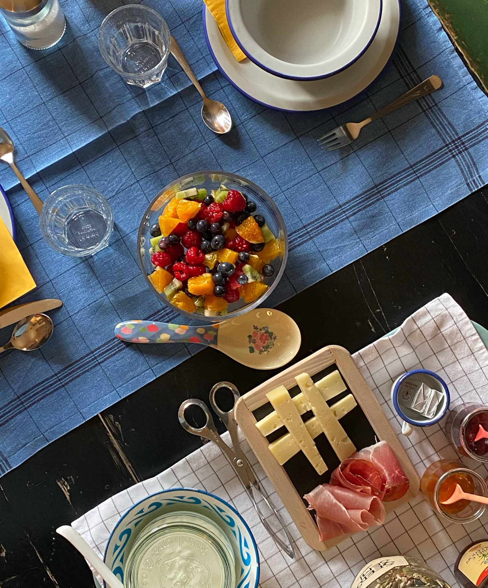 Liebevoll gedeckter Frühstückstisch von oben mit frischem Obstsalat, Käse, Schinken und Marmeladen in rustikalem Geschirr