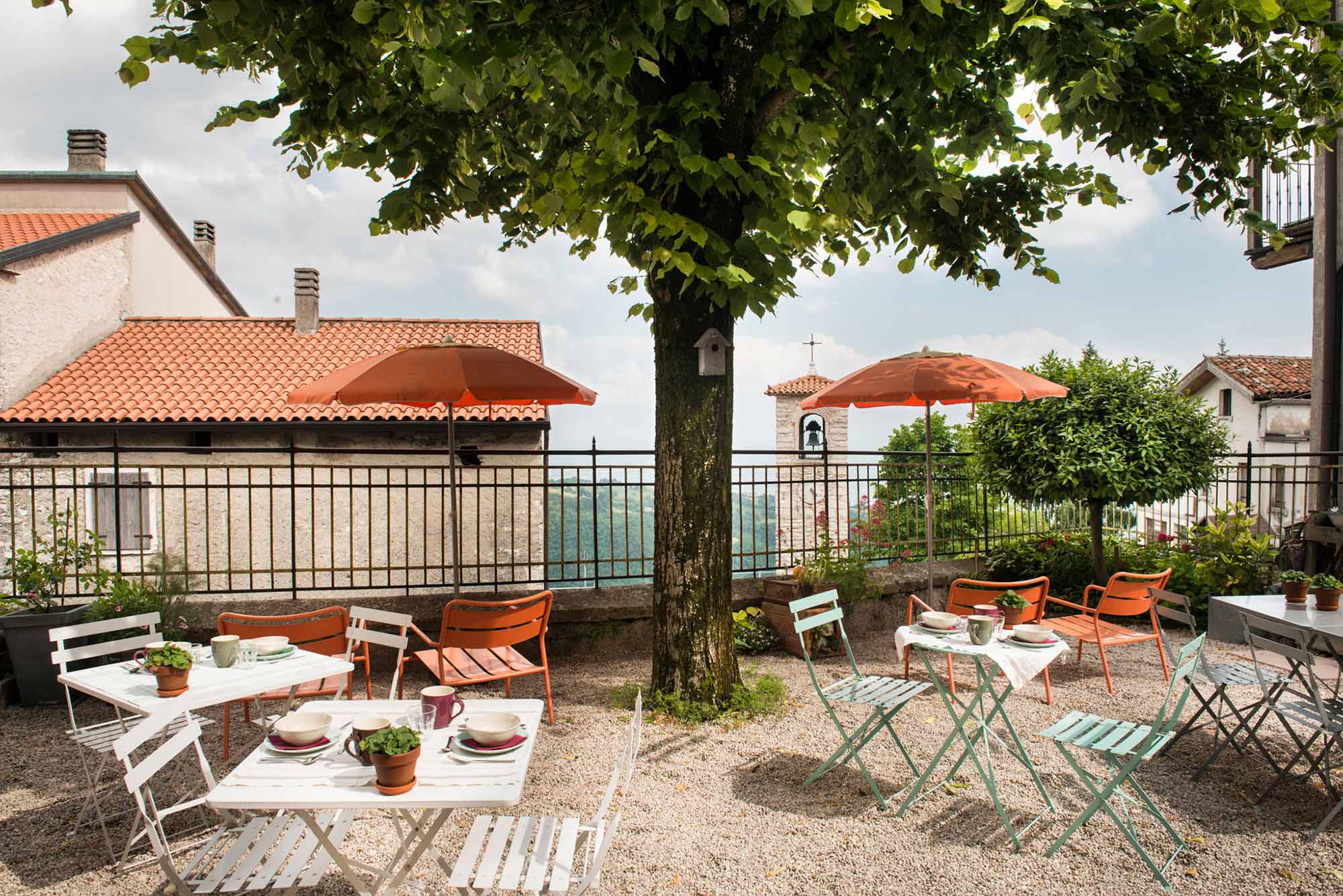 Sonnige Außenterrasse des B&B La Scuola mit bunten Bistro-Tischen, orangefarbenen Sonnenschirmen und Blick auf die Hügellandschaft Venetiens