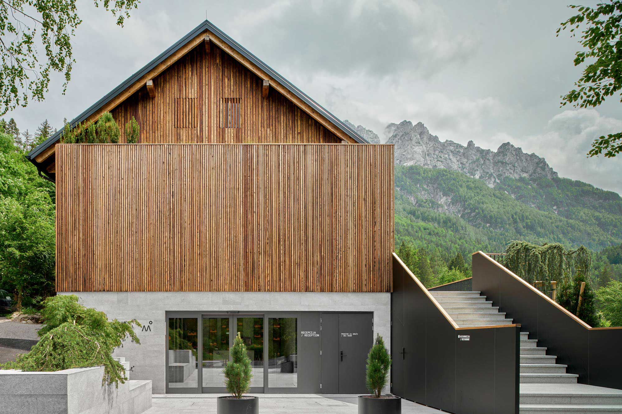 Eingangsbereich des Boutique Hotels Milka in Kranjska Gora mit vertikaler Holzlamellenfassade, Betonunterbau, Rezeptionsschild und Bergkulisse der Julischen Alpen