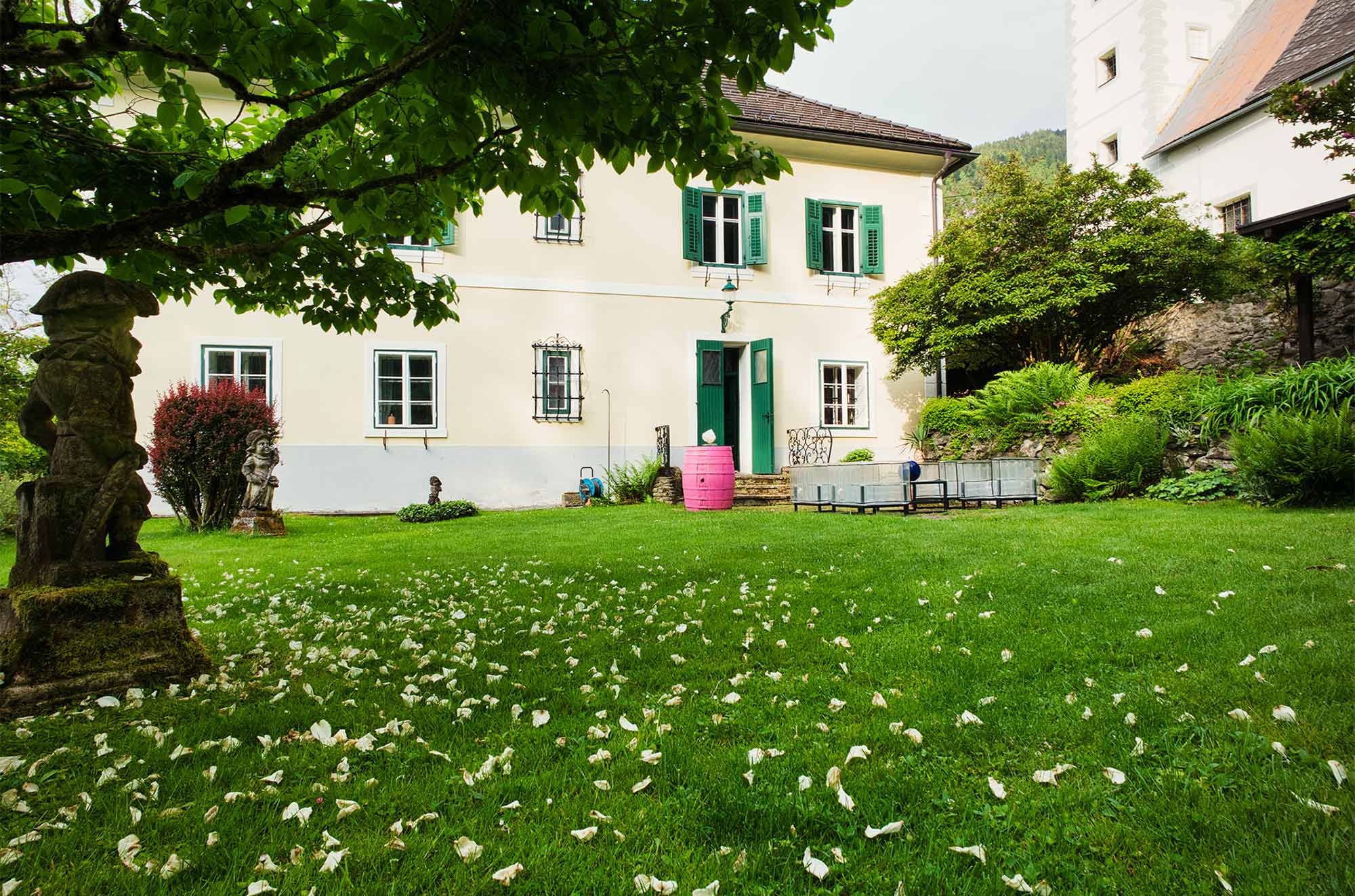 Cremeweiße Fassade des alten Pfarrhauses mit grünen Fensterläden, gepflegtem Garten und Steinfiguren unter blühendem Baum