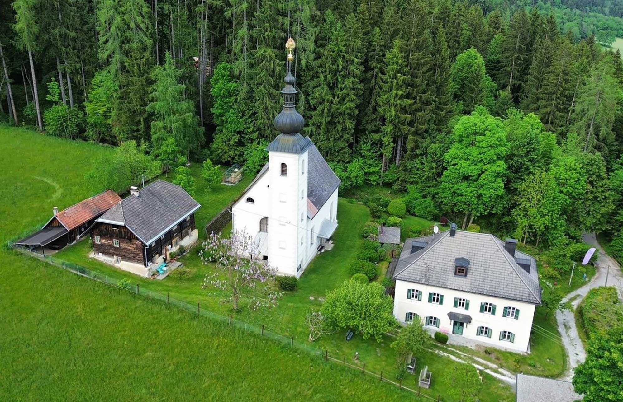 Luftaufnahme des historischen Pfarrhauses „rosa und blau am See" mit Kirche, Nebengebäude und umliegendem Wald in Bodensdorf, Kärnten