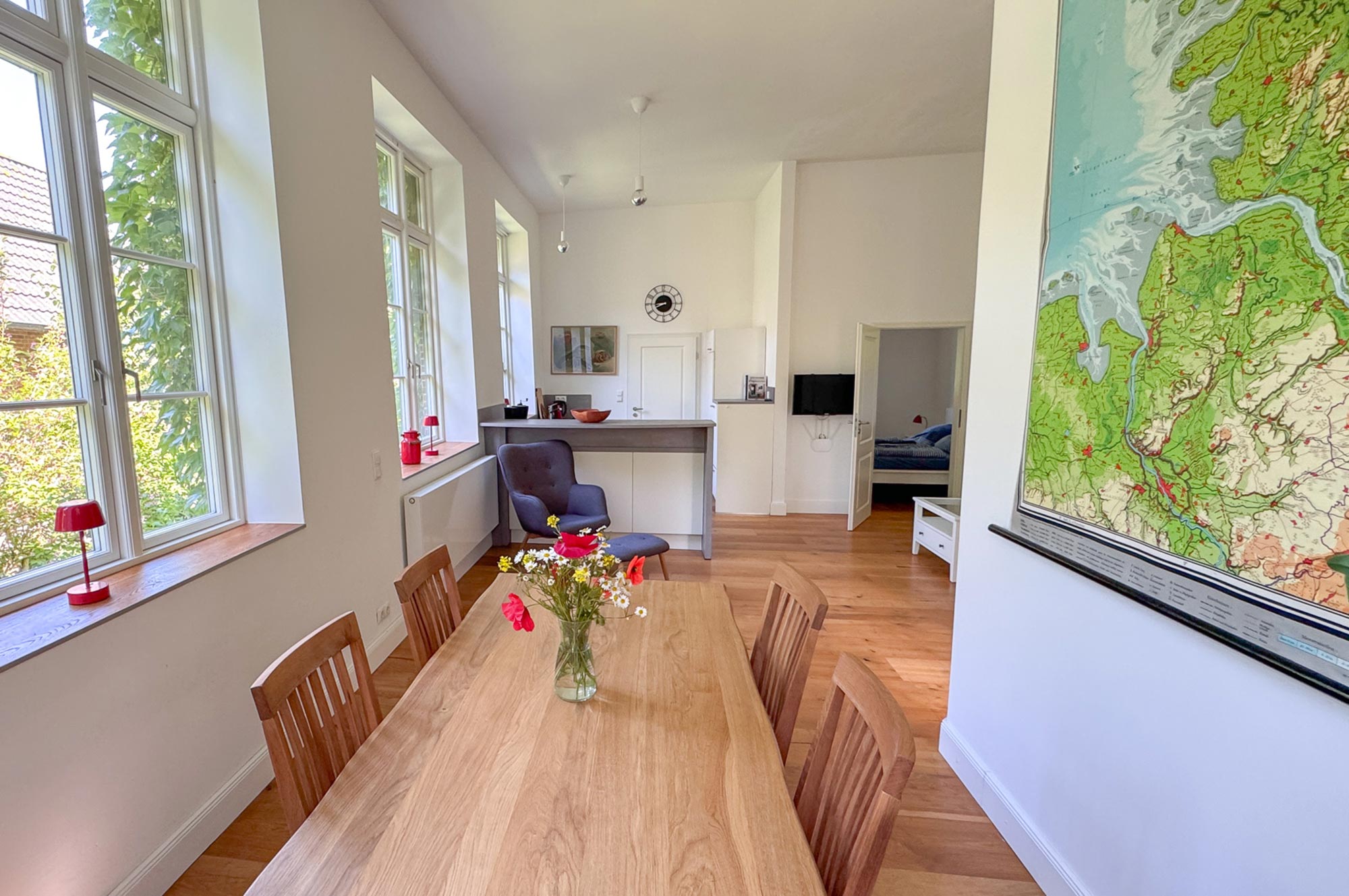 Lichtdurchfluteter Wohn- und Essbereich mit langem Holztisch, gemütlichem Sessel, offener Küche und Blick ins Schlafzimmer