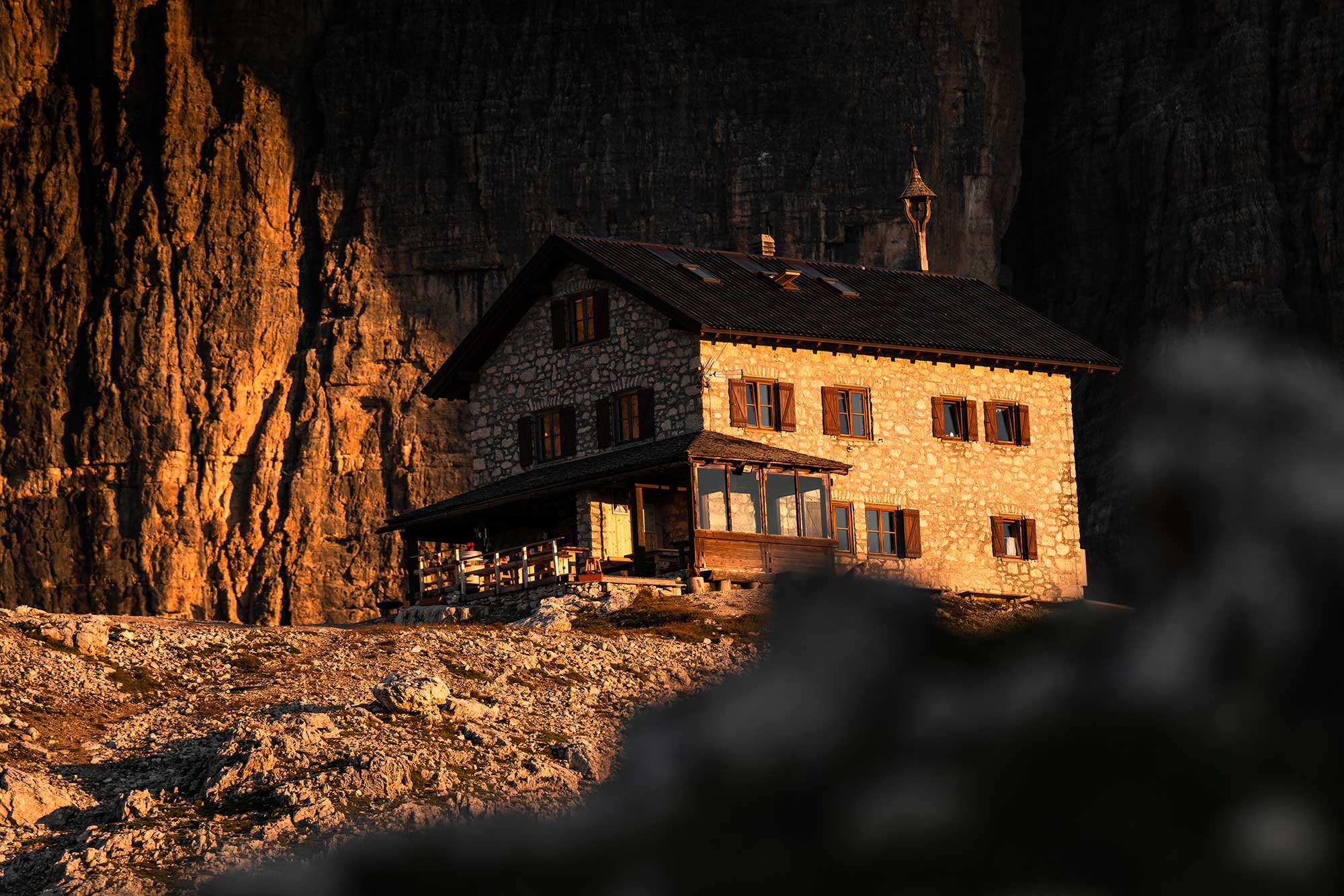 Franz-Kostner-Hütte in den Dolomiten im warmen Abendlicht vor dunkler Felswand der Sellagruppe, historisches Steingebäude mit Holzläden und Veranda