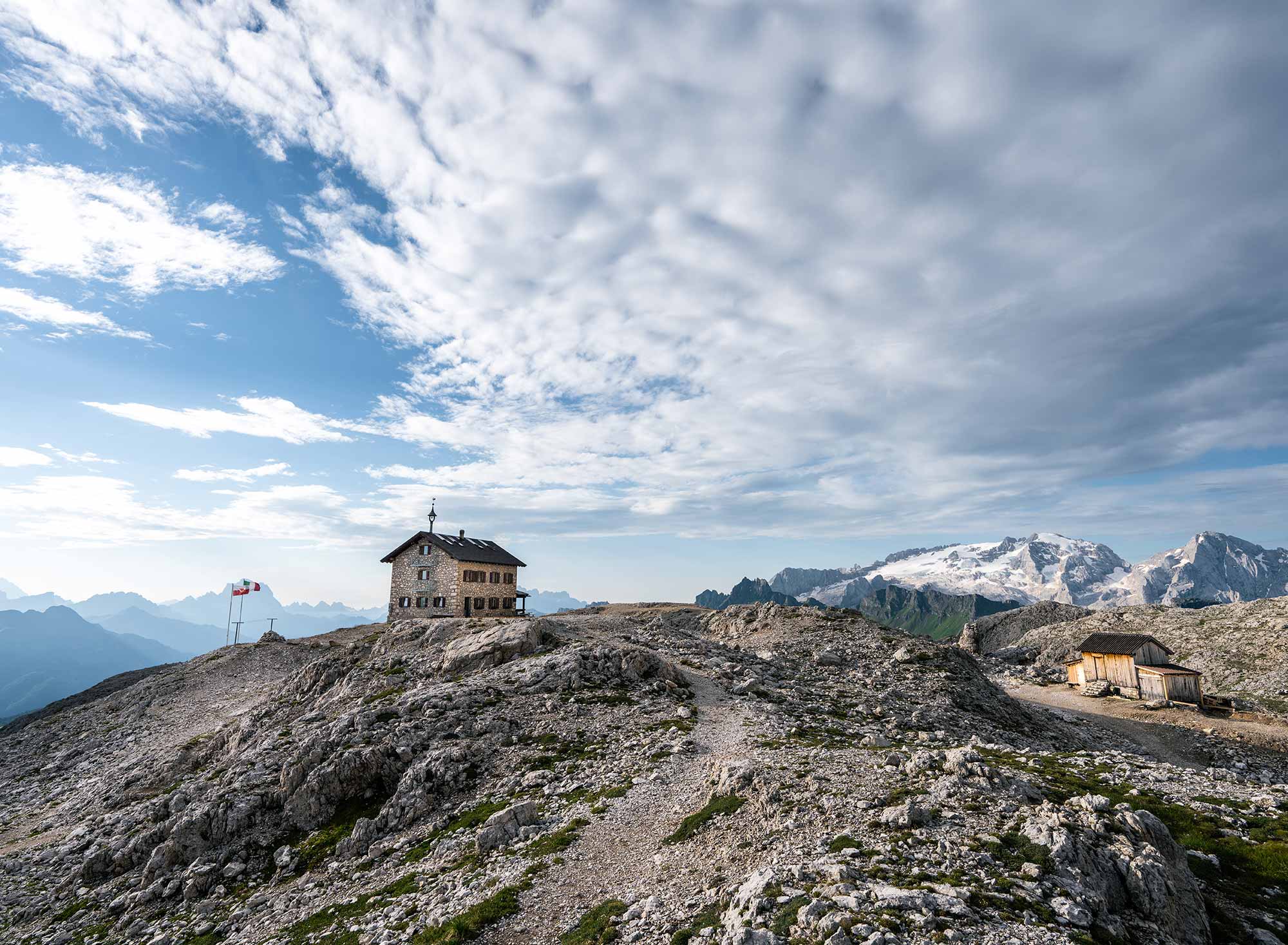Franz-Kostner-Hütte auf felsigem Hochplateau in den Dolomiten mit Wanderweg, Fahnen und Blick auf die schneebedeckte Marmolada unter bewölktem Himmel
