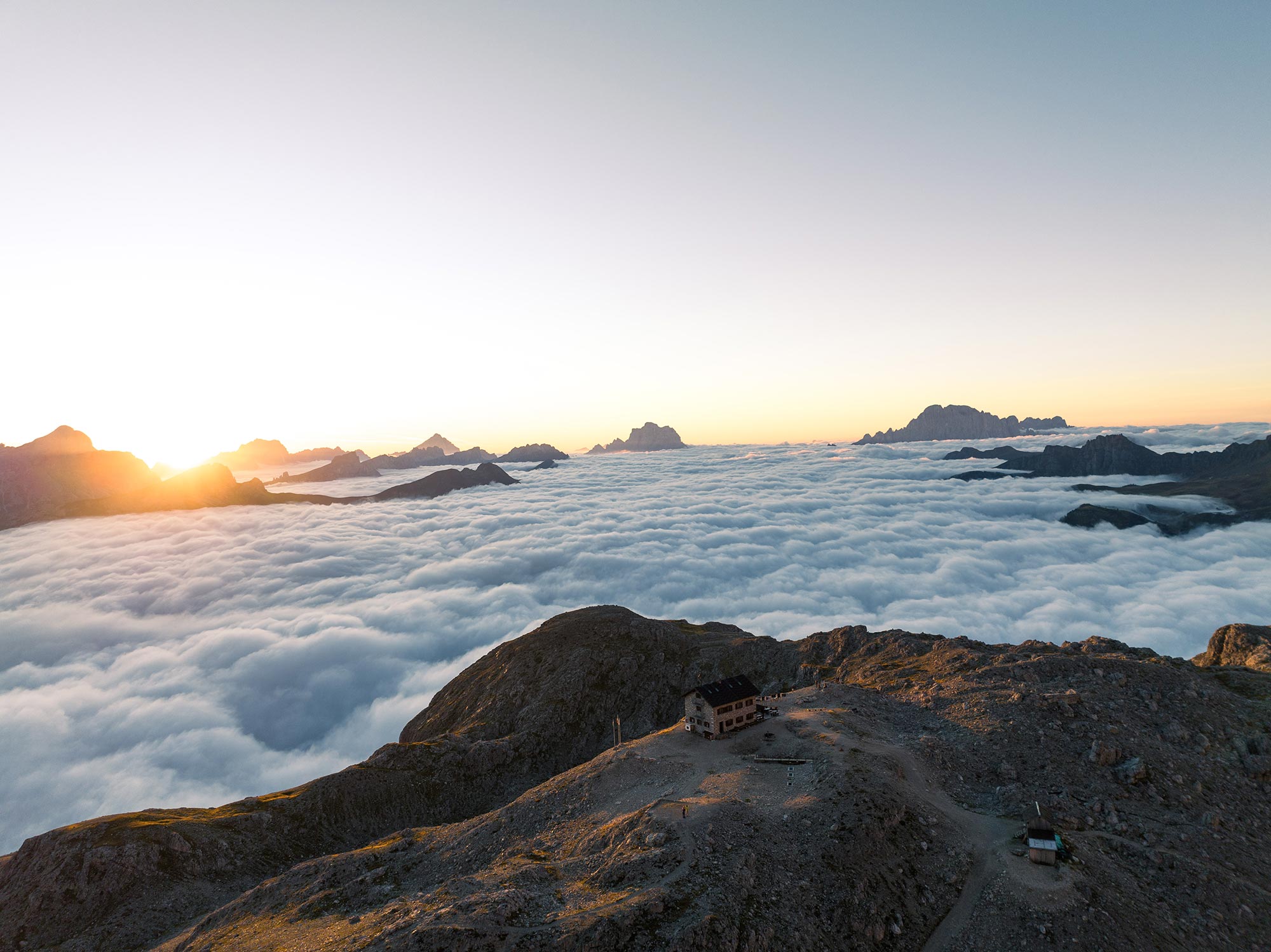 Luftaufnahme der Franz-Kostner-Hütte auf 2.550 Metern in den Dolomiten bei Sonnenaufgang, umgeben von einem weißen Wolkenmeer mit Bergspitzen am Horizont