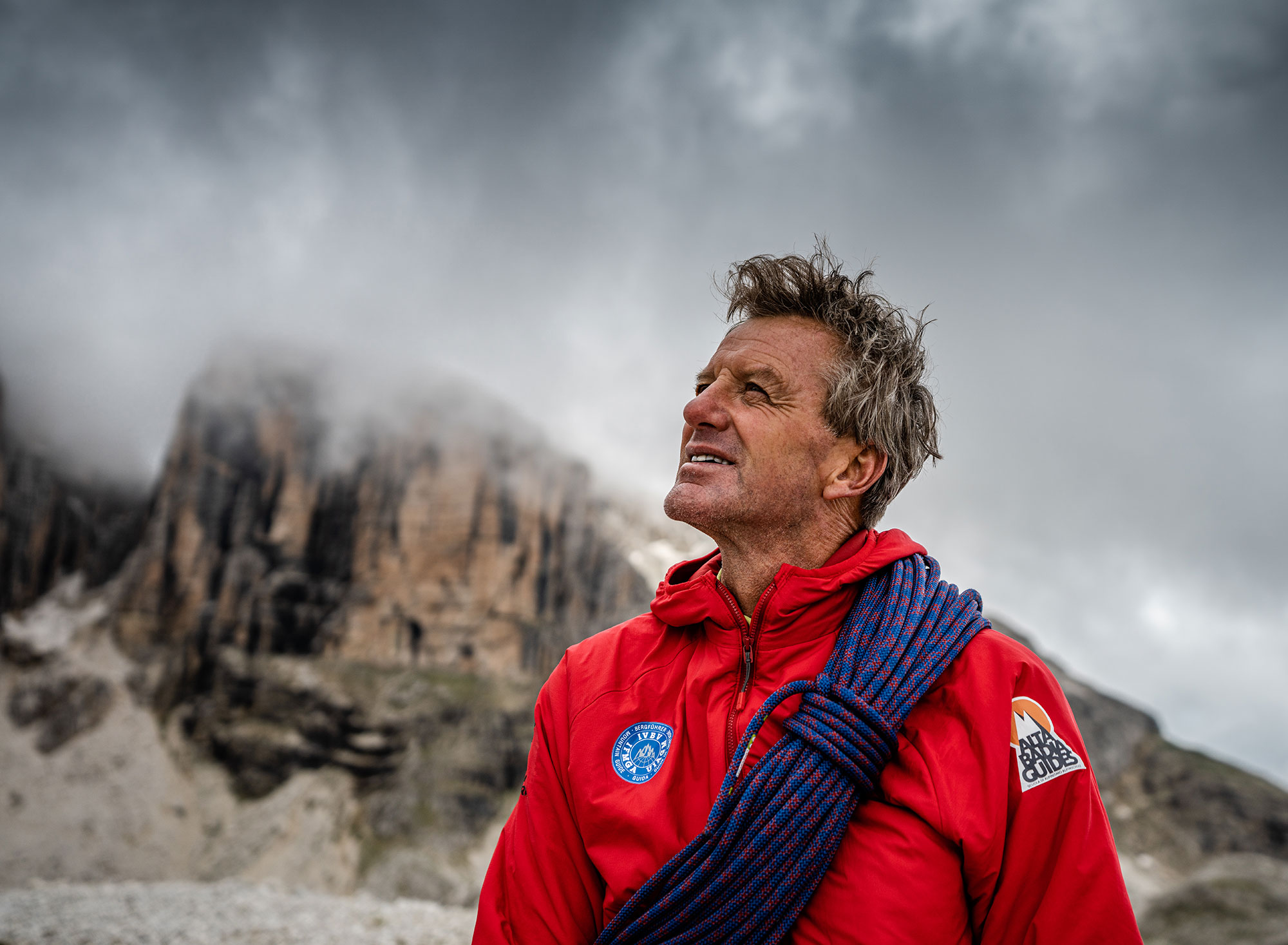 Porträt von Manuel Agreiter als Bergführer mit blauem Kletterseil über der Schulter, roter Jacke mit Alta Badia Guides-Patch, vor bewölktem Dolomitenhimmel