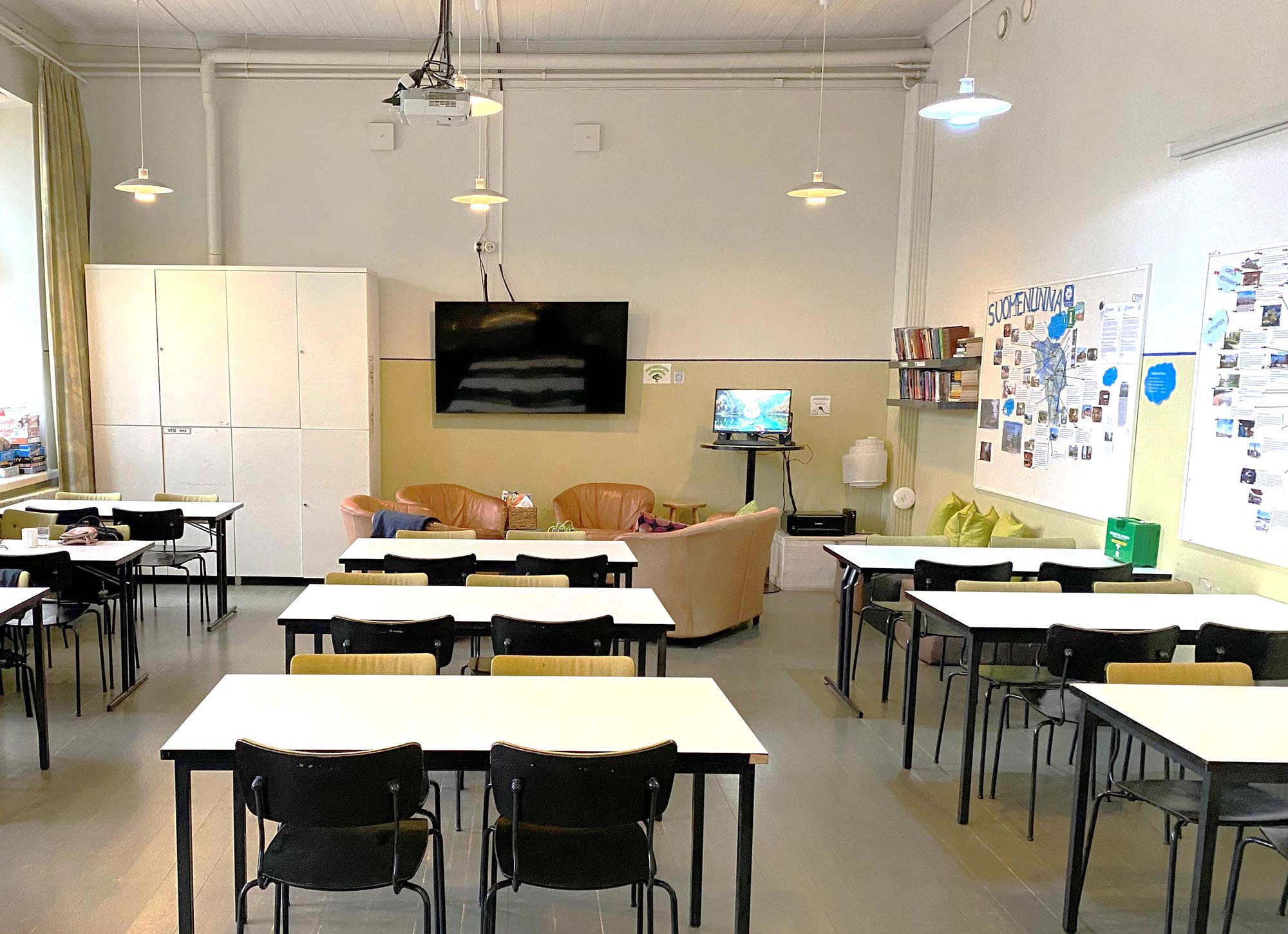 Geräumiger Gemeinschaftsraum im Hostel Suomenlinna mit Tischreihen, Sitzecke, Bücherregal und Informationswänden über die Insel