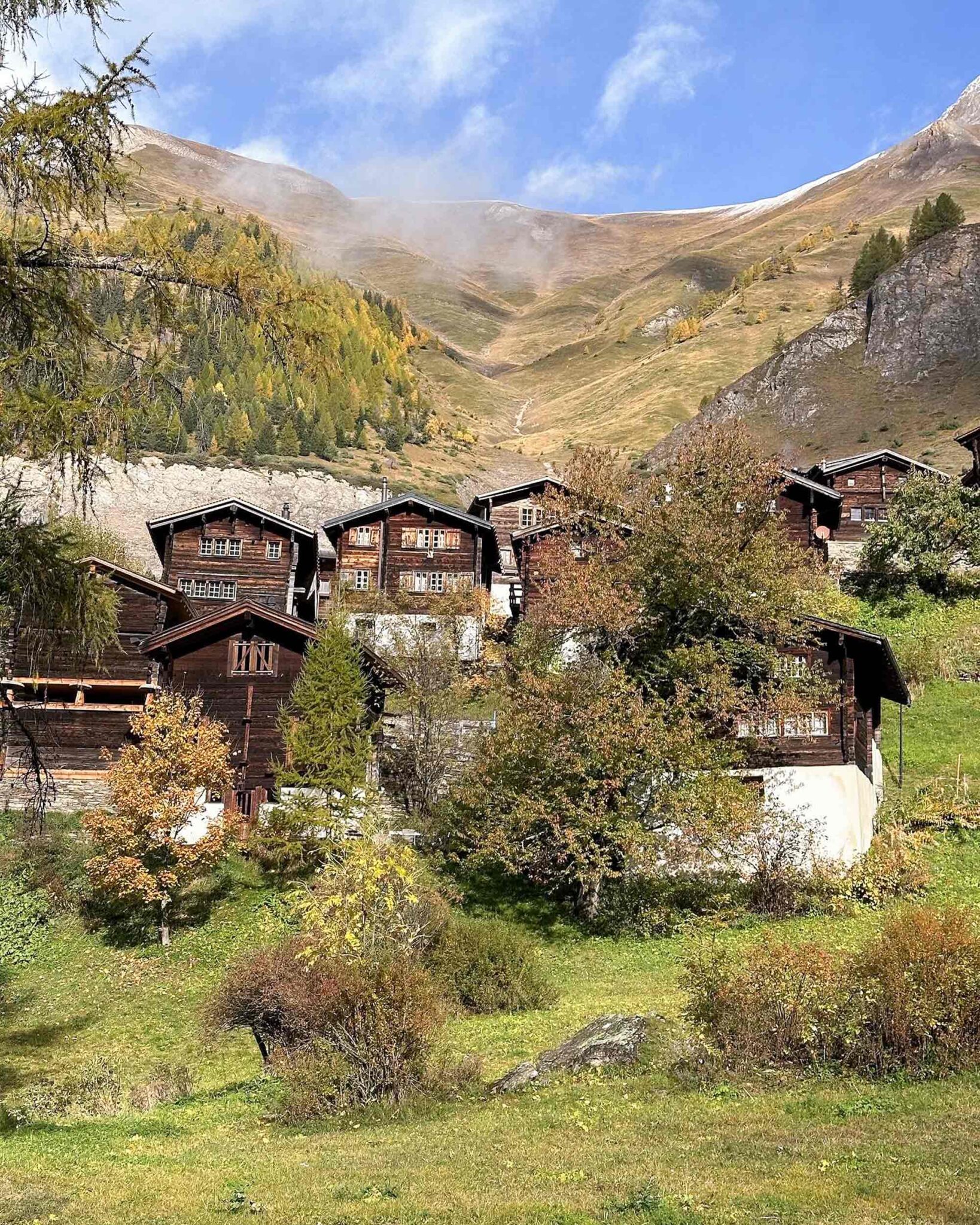 Blick auf das Walliser Bergdorf Martisberg im Herbst: traditionelle dunkle Holzhäuser eingebettet in eine weite Berglandschaft mit herbstlich verfärbten Lärchen