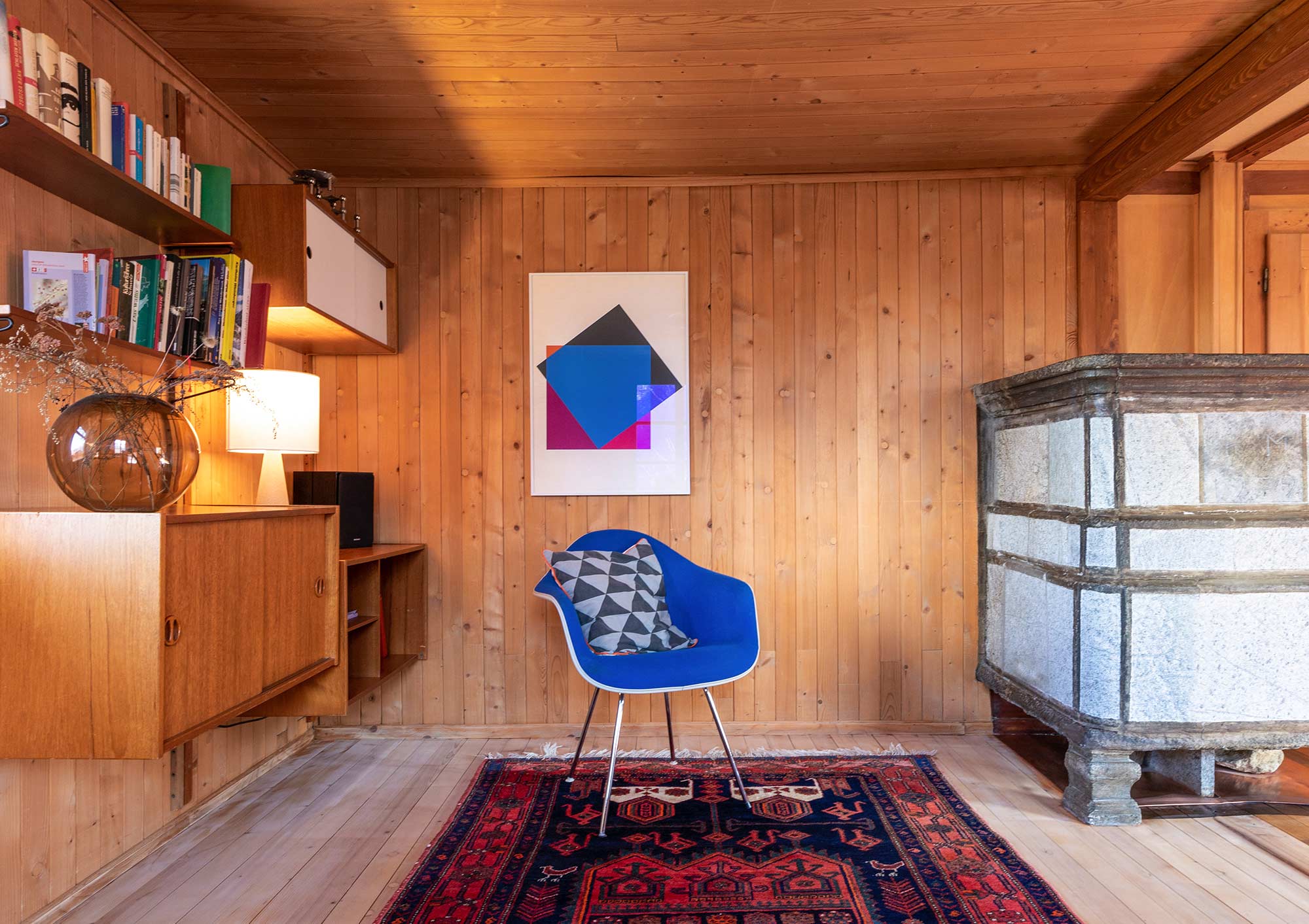 Wohnzimmer im Josihaus in Martisberg mit Holzvertäfelung, historischem Natursteinkachelofen, blauem Designstuhl, Vintagemöbeln und moderner Kunst an der Wand