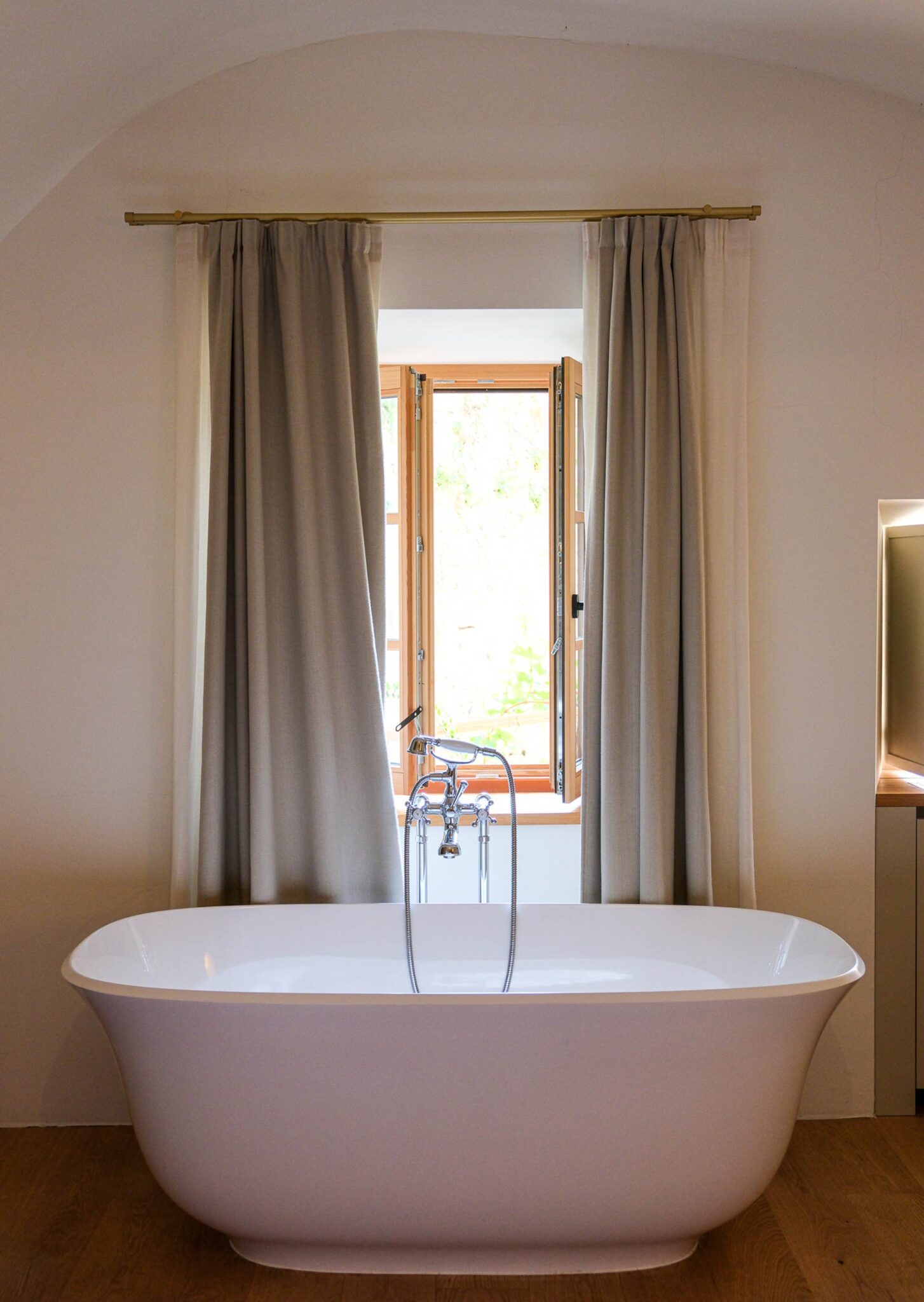 Freistehende weiße Badewanne im Gewölbebad der Pension Leuchtenburg mit geöffnetem Holzfenster und leinengrauen Vorhängen