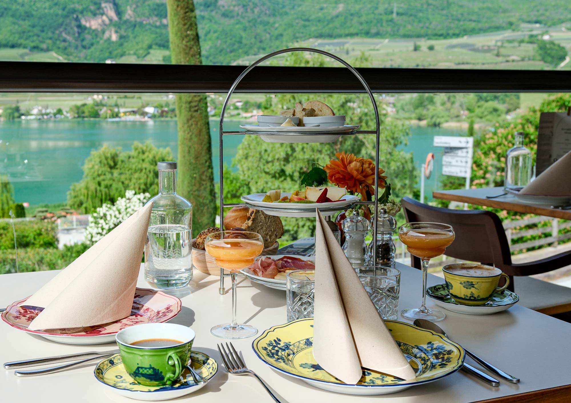Frühstückstisch der Pension Leuchtenburg mit dreistöckiger Etagère, buntem Geschirr und Blick auf den türkisblauen Kalterer See
