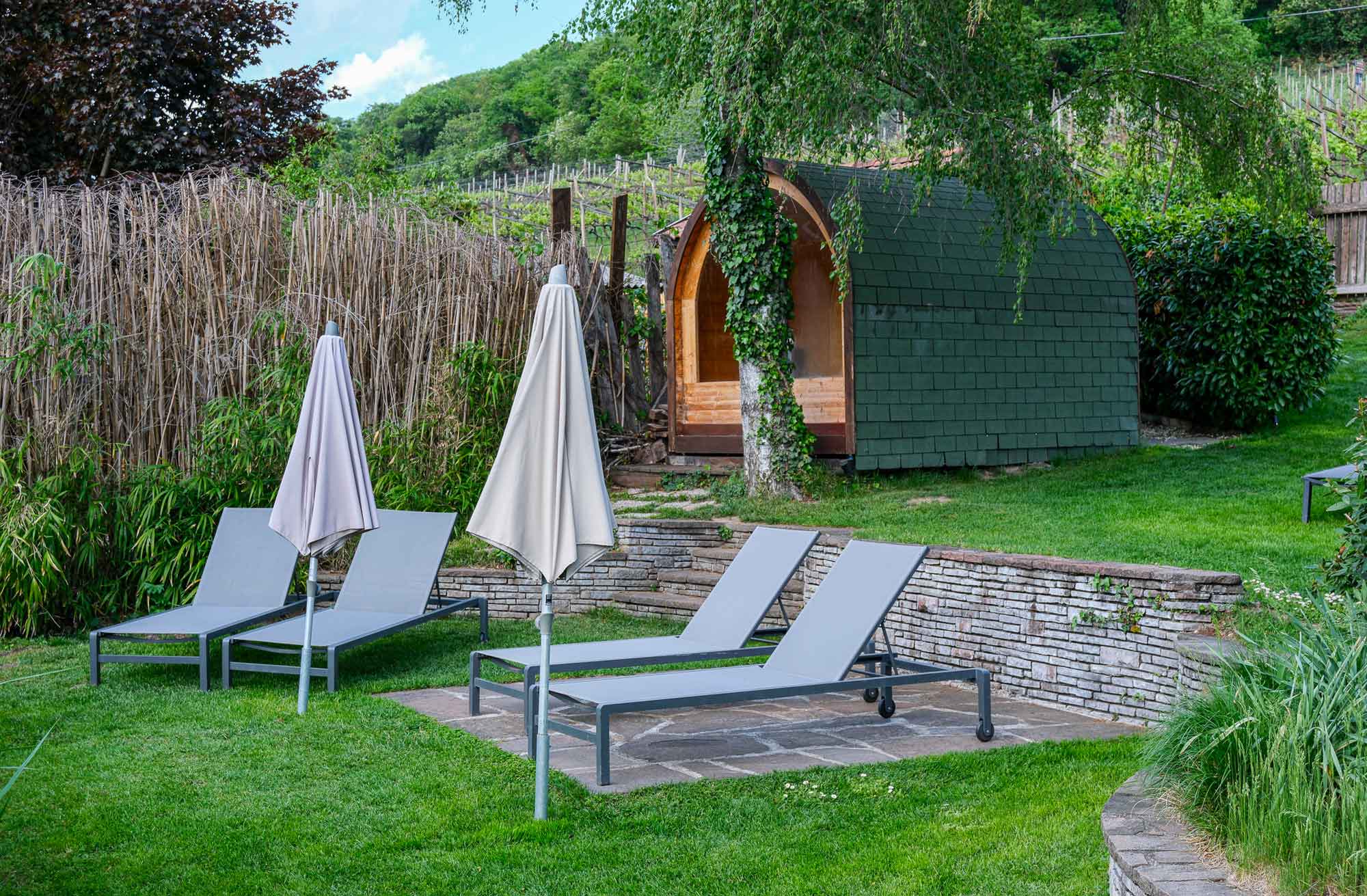 Gartenbereich der Pension Leuchtenburg in Kaltern mit Liegestühlen, Sonnenschirmen und grünem Saunafass zwischen Weinreben