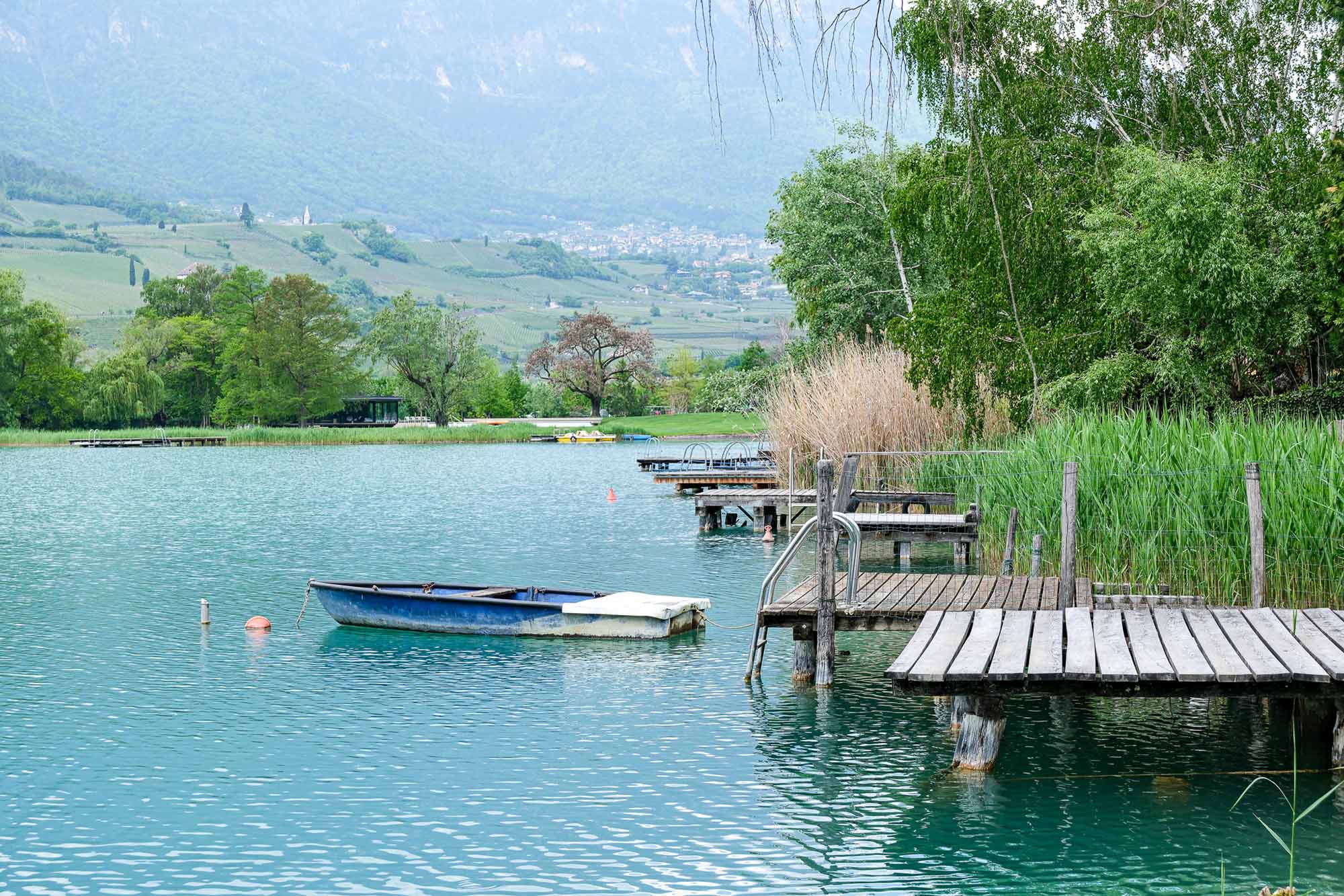 Kalterer See mit Holzsteg, blauem Ruderboot und Blick auf begrünte Weinberge und Berge Südtirols im Frühling