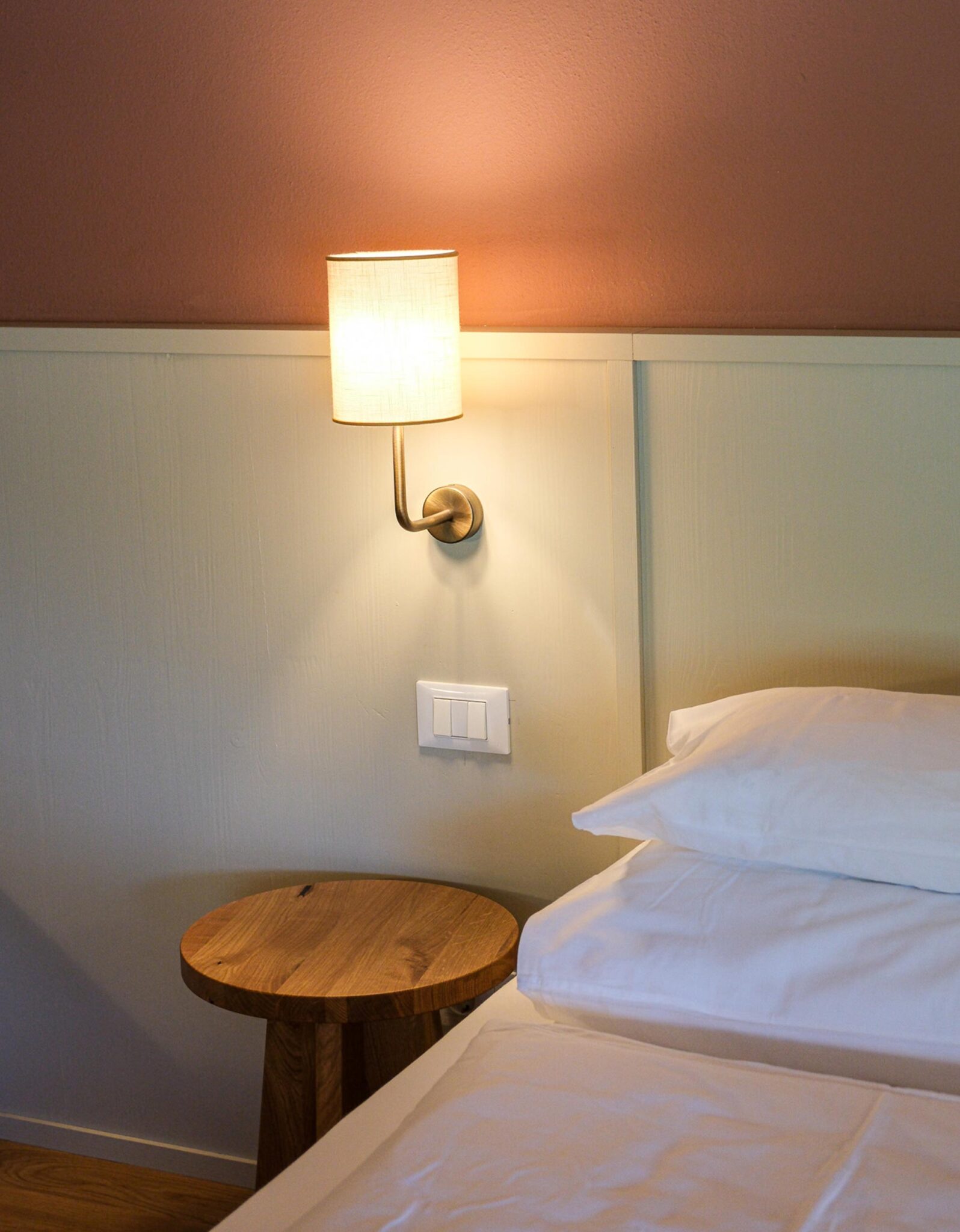 Zimmerdetail der Pension Leuchtenburg mit weißem Bett, rundem Holznachttisch und Messingwandlampe vor terrakottafarbener Wand