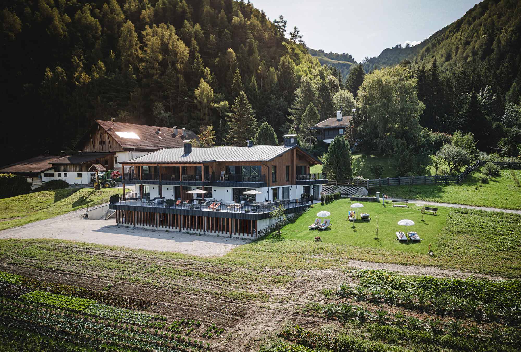 Luftaufnahme des Perntal Retreats in Gais, Südtirol: modernes Landhaus mit großer Sonnenterrasse, Liegewiese, Gemüsebeeten und dem historischen Bauernhof im Hintergrund