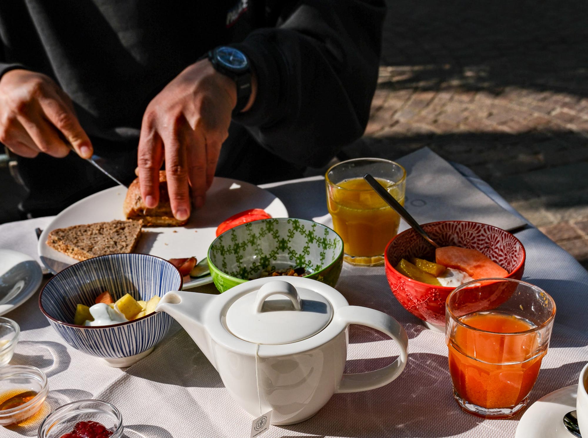 Frühstückstisch am Stroblhof Eppan mit bunten Schälchen, Teekannen, frischen Säften und Vollkornbrot im Freien bei Sonnenlicht