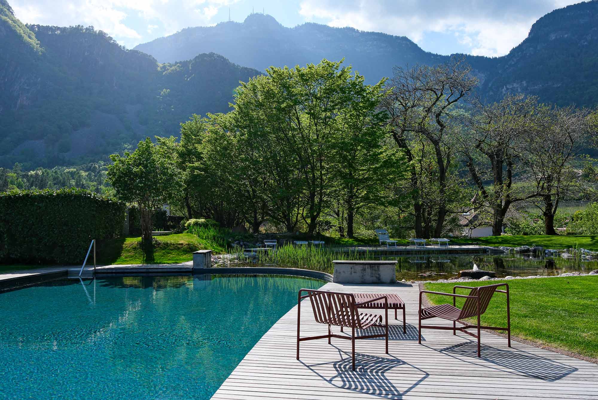 Naturpool des Hotel Stroblhof in Eppan, Südtirol, mit türkisblauem Wasser, rotem Sitzmöbel auf Holzdeck und grünem Bergpanorama im Hintergrund