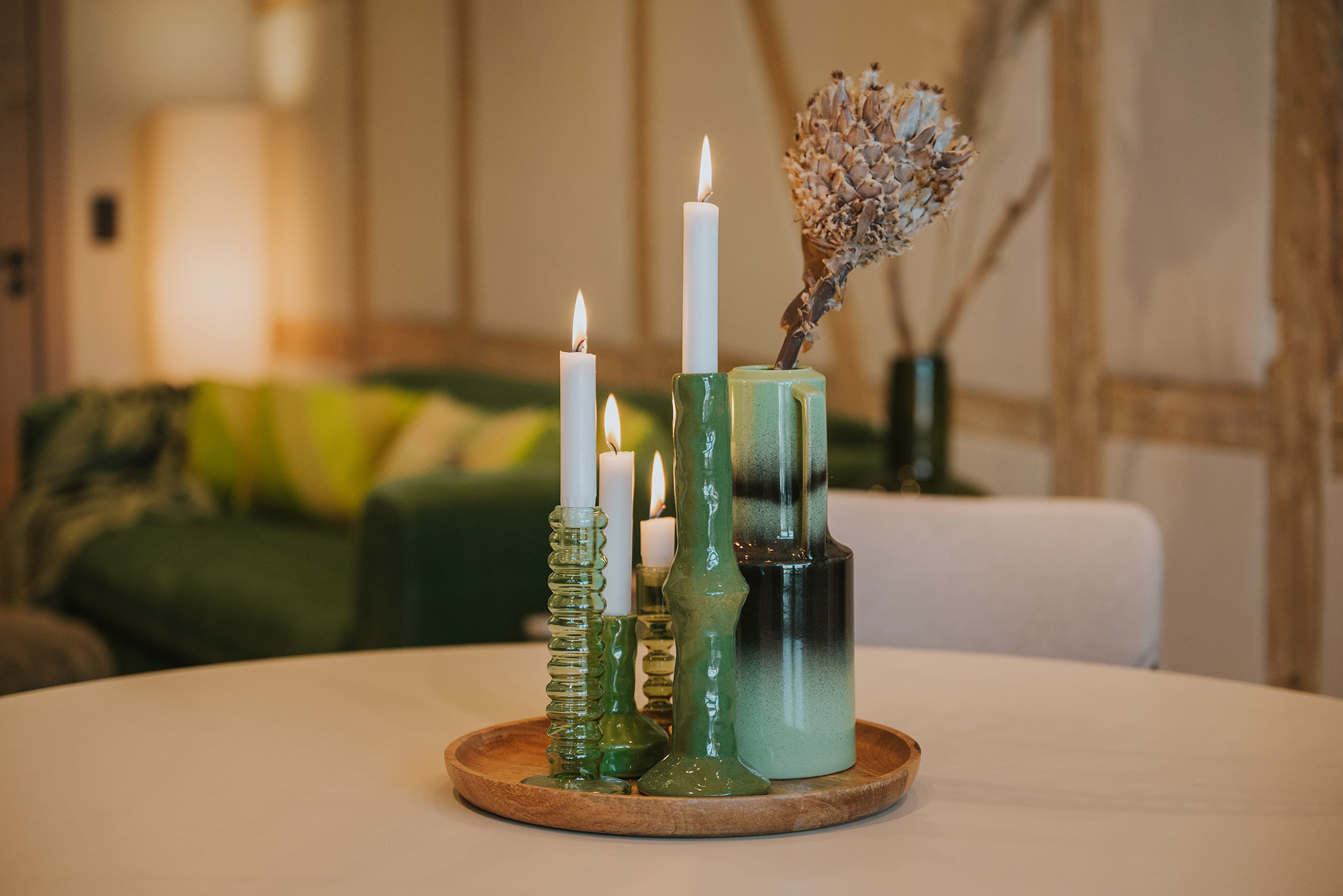 Stimmungsvolles Detailfoto mit brennenden Kerzen, grünen Keramikvasen und Trockenblumen auf Holzteller, im Hintergrund grünes Sofa