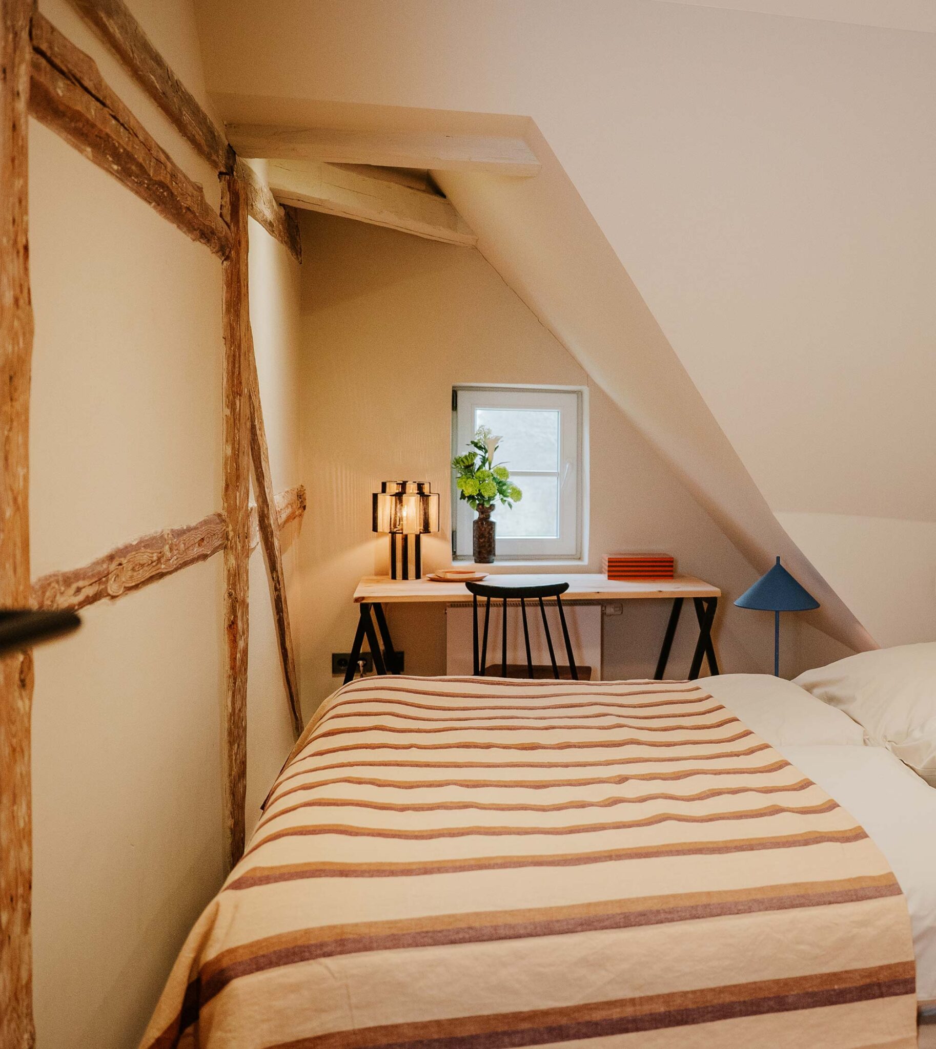 Gemütliches Schlafzimmer im Dachgeschoss mit sichtbarem Fachwerk, gestreifter Tagesdecke und kleinem Schreibtisch am Fenster