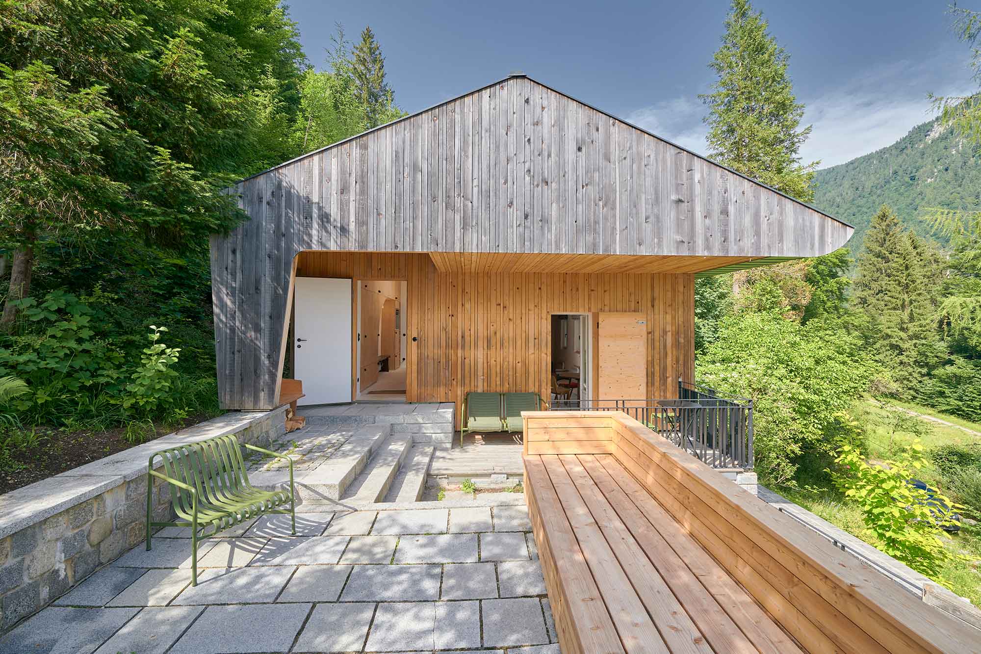 Außenansicht des Ferienhauses Urfeld 26 am Walchensee mit verwitterter Holzfassade, überdachter Terrasse mit grünen Stühlen, Holzdeck und Blick in die bewaldete Berglandschaft