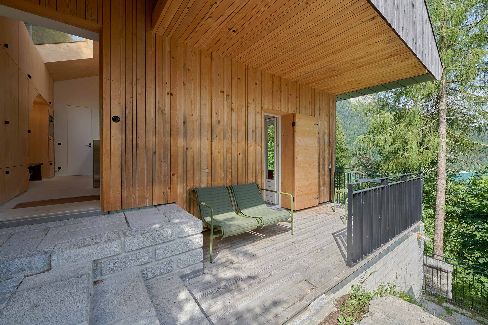 Überdachte Holzterrasse des Ferienhauses Urfeld 26 mit zwei grünen Liegestühlen, Blick auf den Walchensee und die umliegenden Berge im Sommer