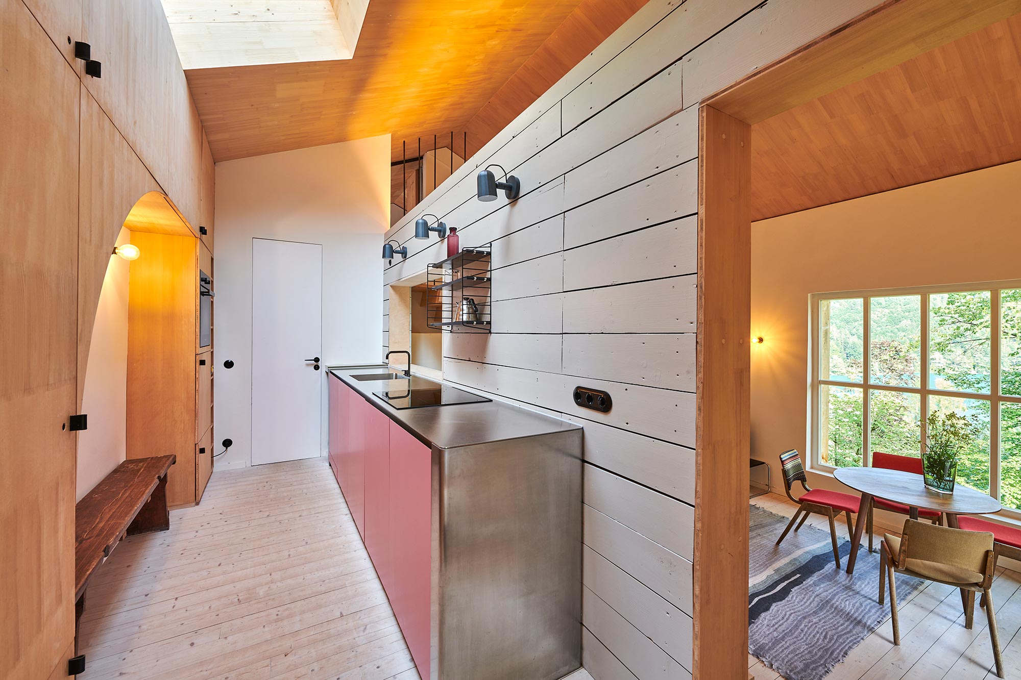Offene Küche im Ferienhaus Urfeld 26 mit rosa Küchenfronten, Edelstahloberfläche, Holzverkleidung und Oberlicht sowie Blick in den Essbereich mit Seefenster