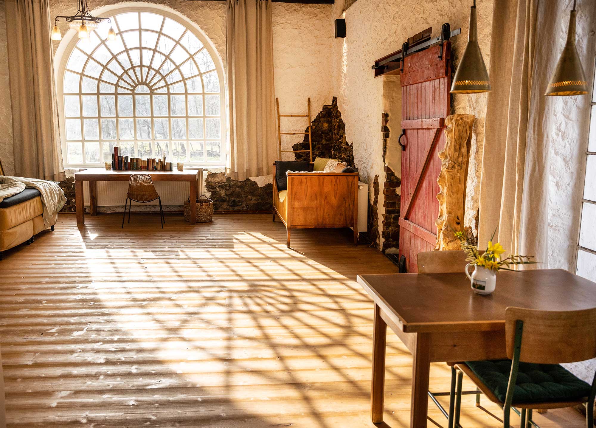Lichtdurchflutetes Zimmer im Ferienhaus Gut Neuwerk mit großem Rundbogenfenster, Holzboden und rustikaler Einrichtung