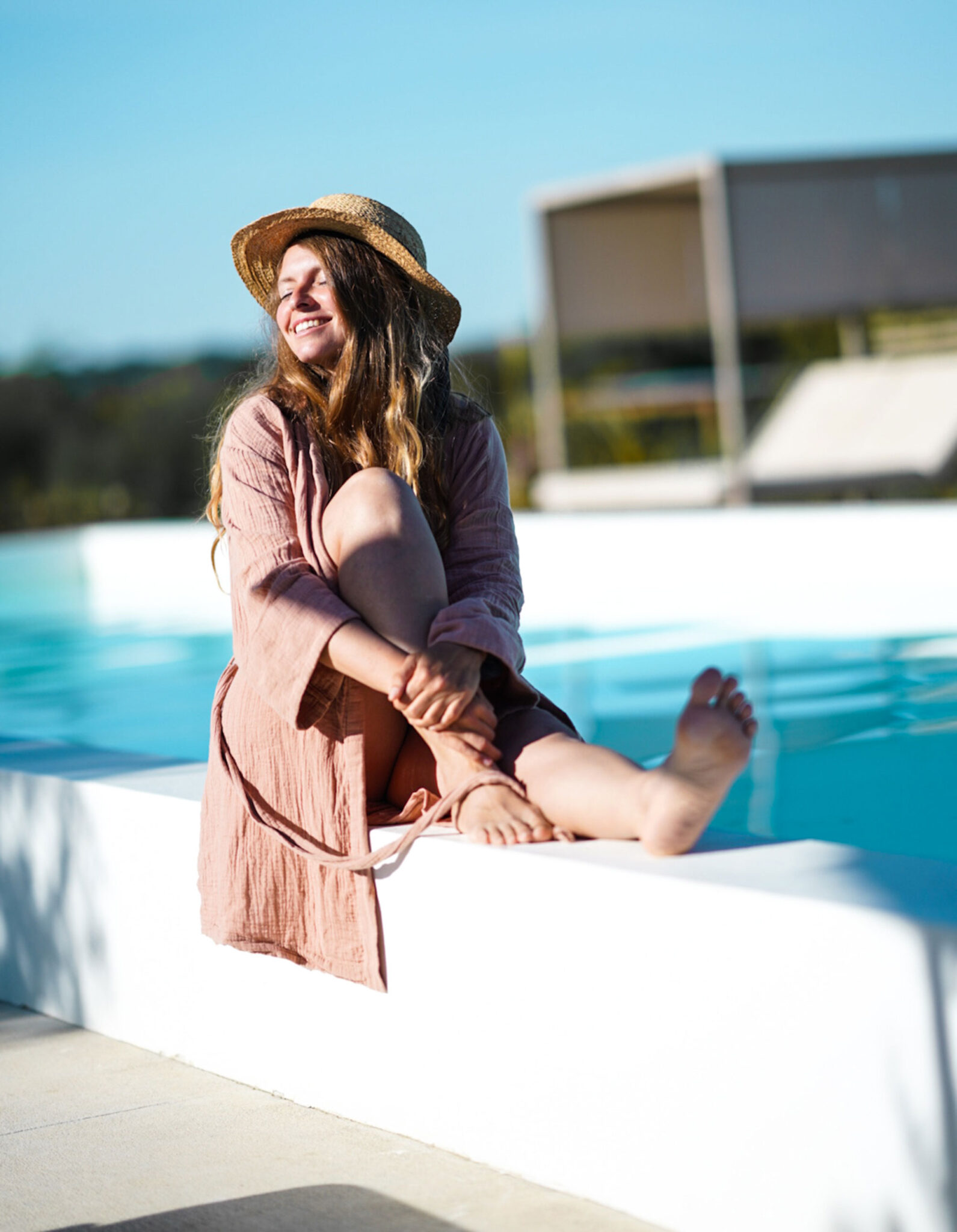 Frau in rosafarbenem Leinenkleid und Strohhut sitzt lachend am Rand des Infinity-Pools im Gandum Village