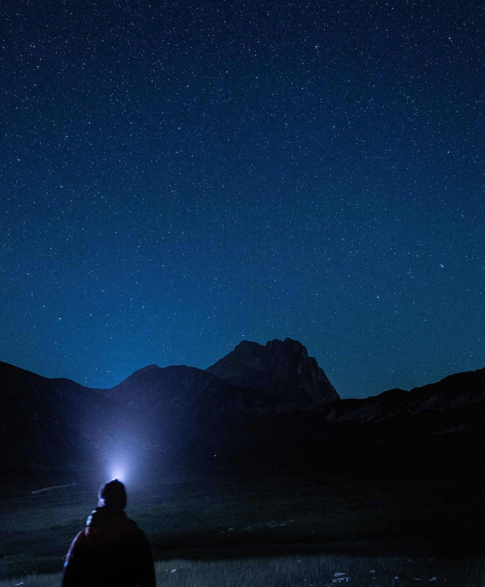 Person mit Stirnlampe blickt in den Sternenhimmel über einer Bergsilhouette