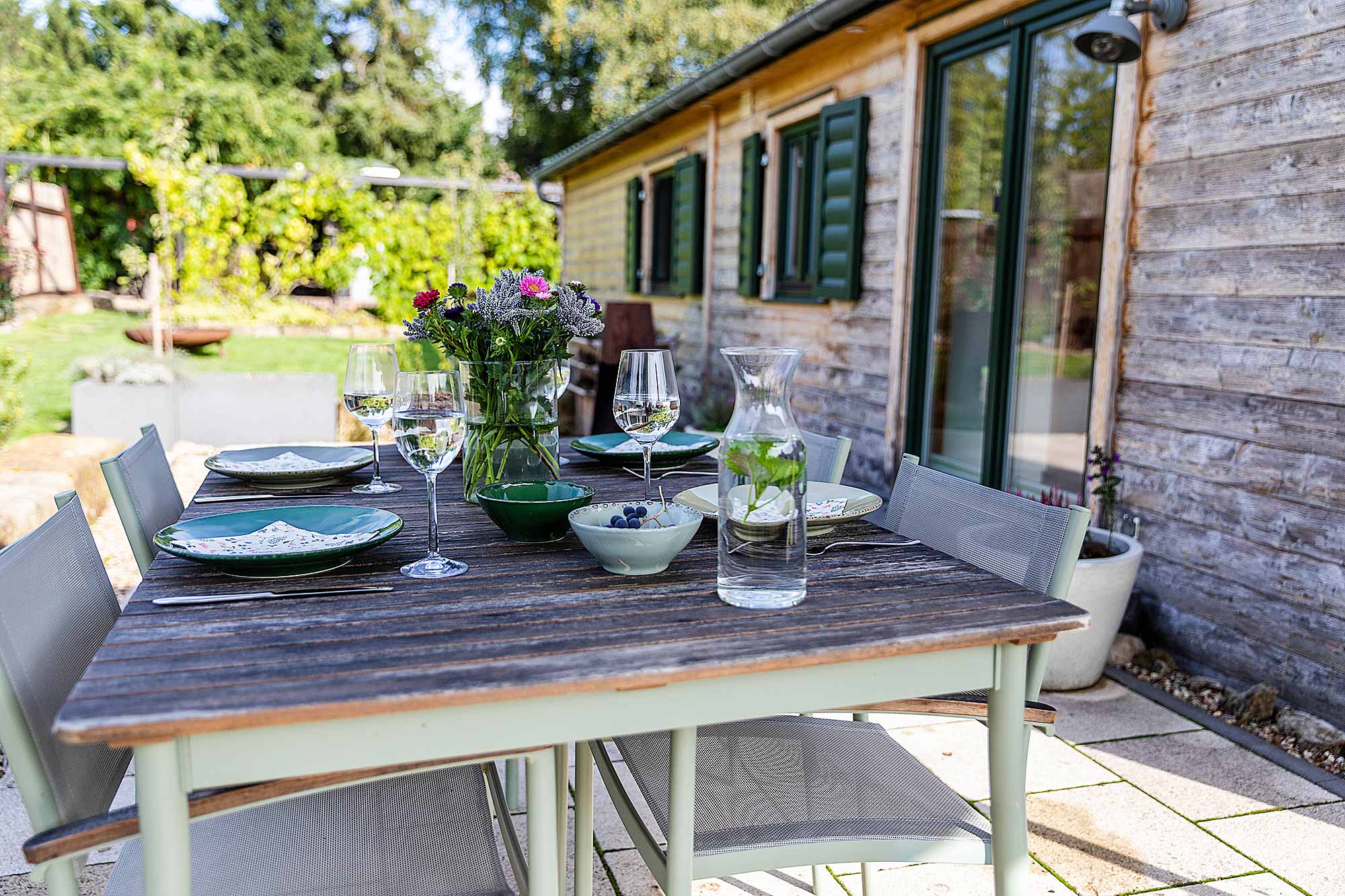 Gedeckter Tisch mit Geschirr, Weingläsern und Blumenstrauß auf der Terrasse vor dem Ferienhaus Waldhauszeit