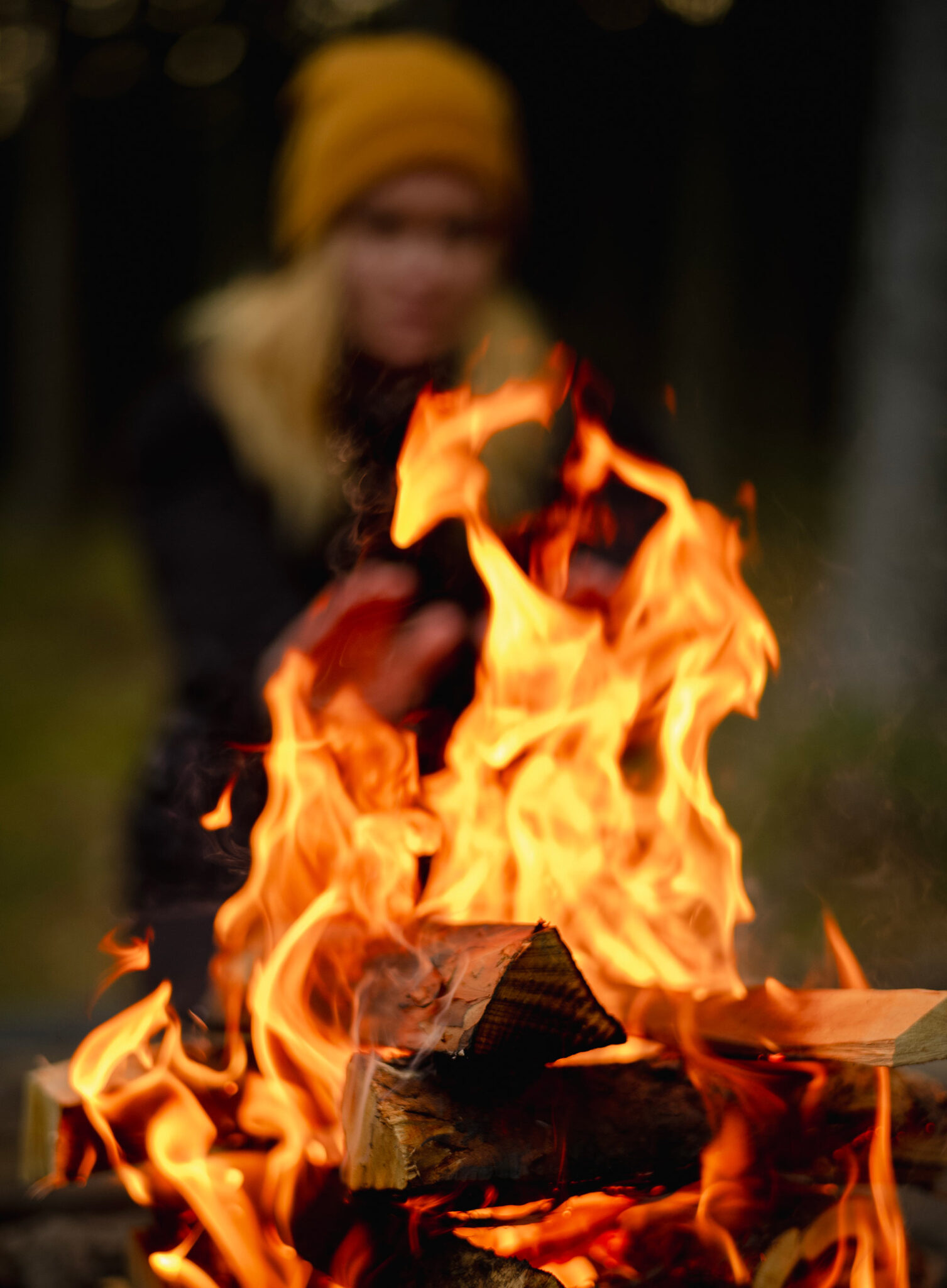 Nahaufnahme eines Lagerfeuers, im Hintergrund unscharf eine Person mit gelber Mütze beim Wärmen der Hände