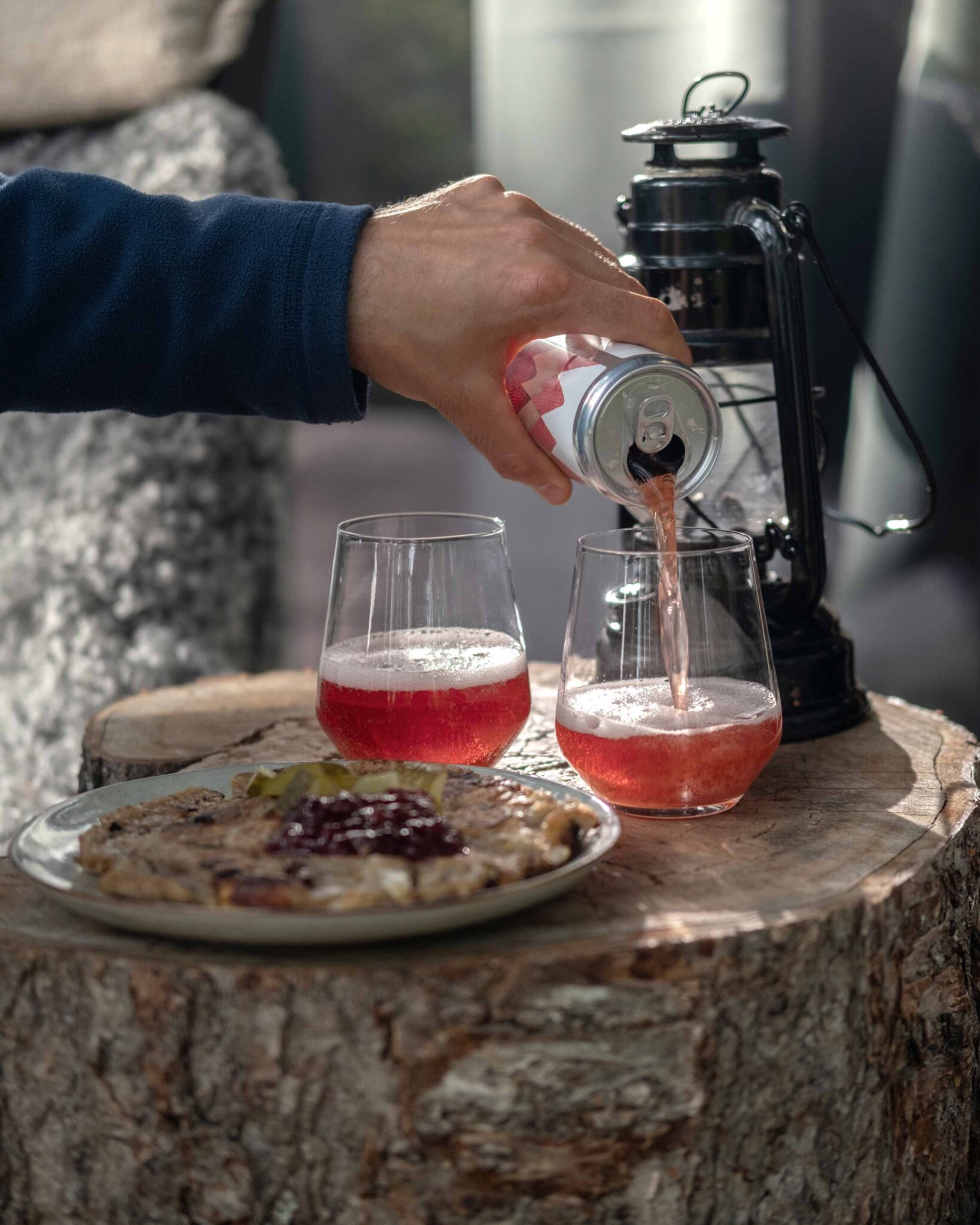 Hand gießt ein Getränk aus der Dose in zwei Gläser, daneben ein Teller mit herzhaftem Abendessen und Preiselbeermarmelade auf einem Baumstamm-Tisch