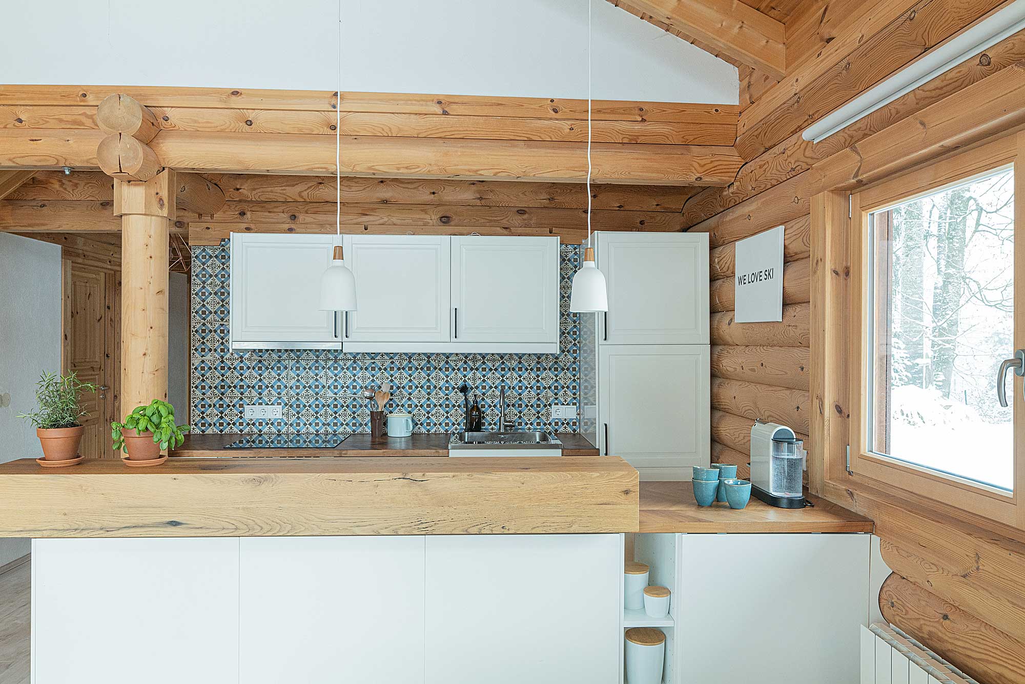 Offene Küche im Blockhaus Hütte am Wald mit weißen Schränken, blau gemusterten Fliesen und Blick auf den verschneiten Wald