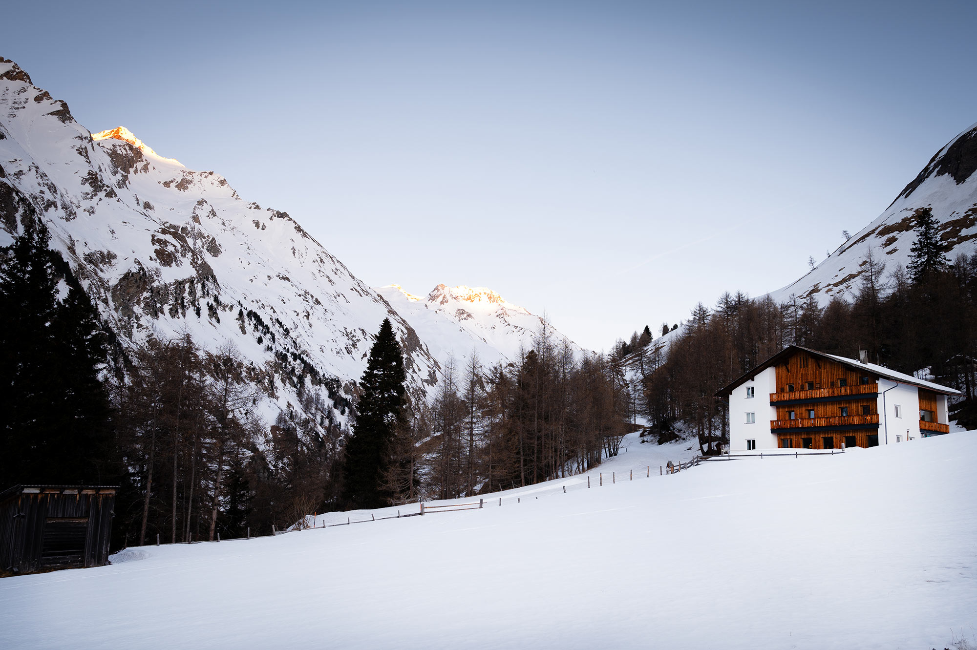 Außenansicht der MoaAlm Mountain Retreat im Schnee bei Abenddämmerung mit schneebedeckten Gipfeln im Alpenglühen
