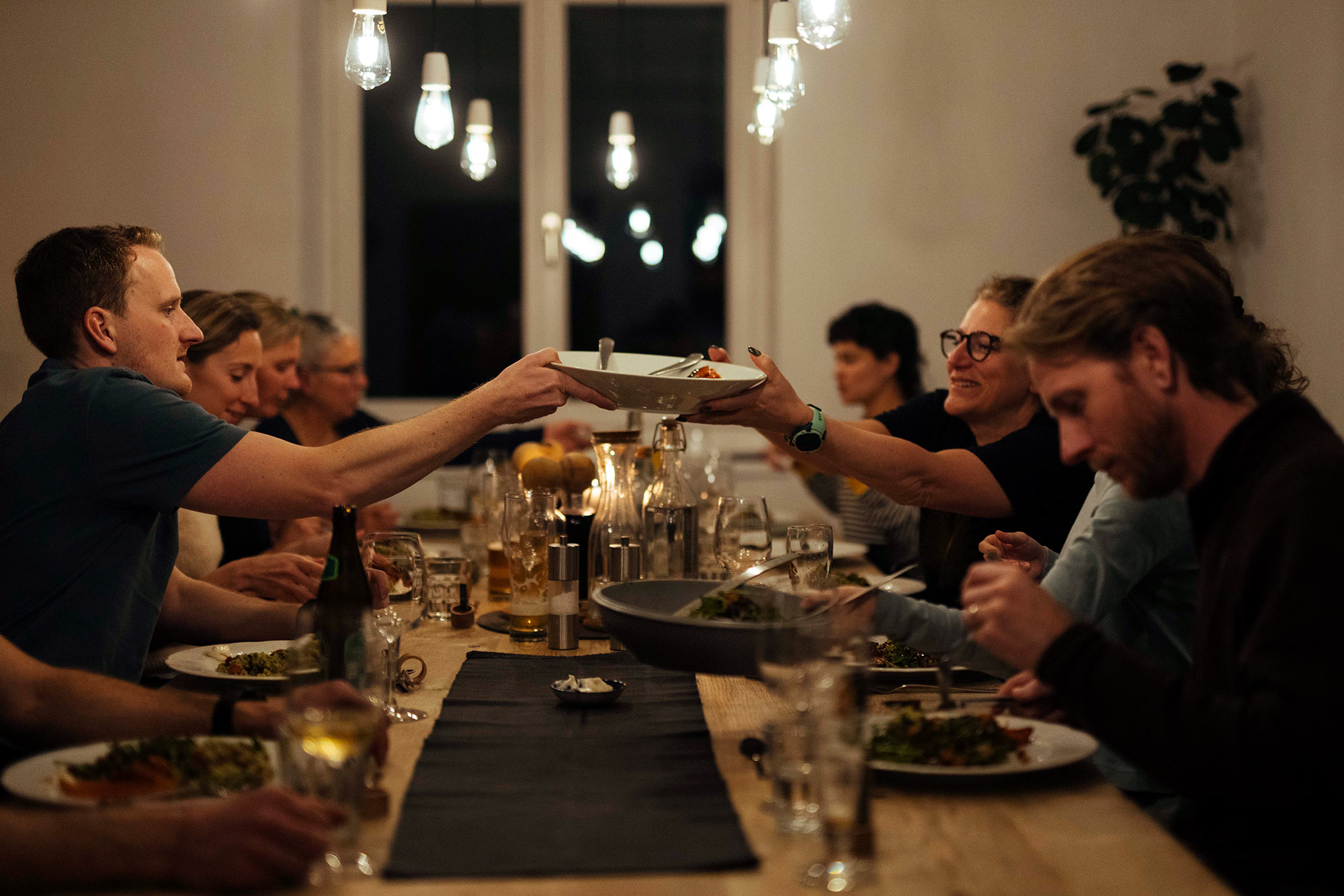 Gäste der MoaAlm Mountain Retreat sitzen abends gemeinsam am langen Tisch und reichen sich Teller mit veganem Essen