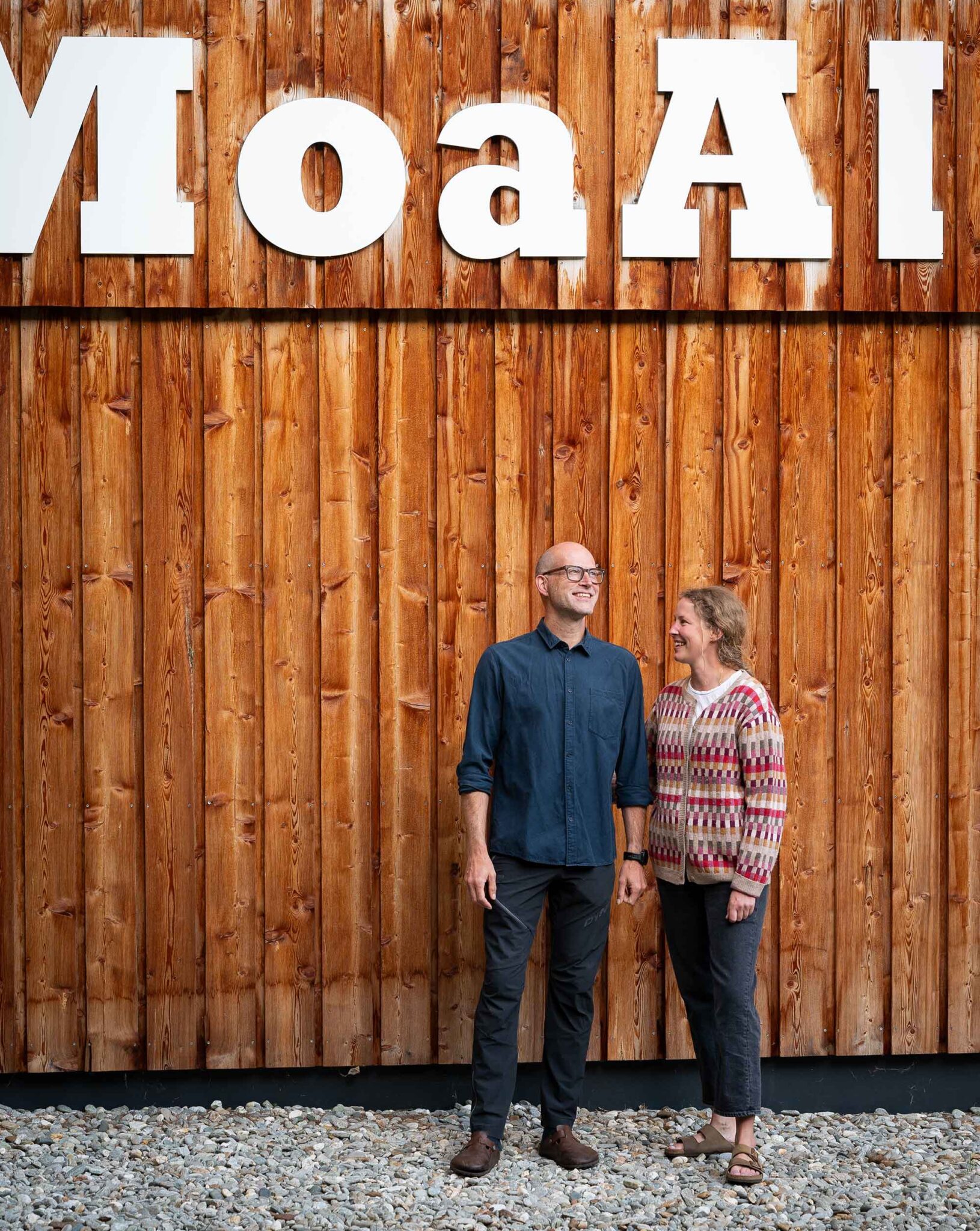 Sophie und Tobias, die Gastgeber der MoaAlm Mountain Retreat, stehen lachend vor der Holzfassade mit MoaAlm-Schriftzug