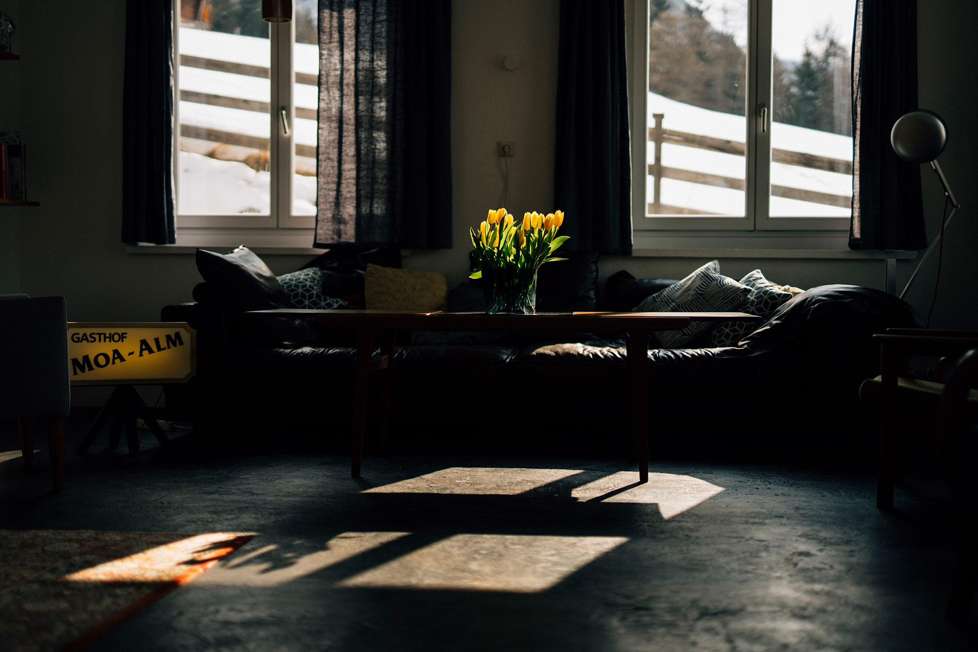 Gemütliches Wohnzimmer der MoaAlm Mountain Retreat mit dunklem Ledersofa, gelben Tulpen und dem alten „Gasthof Moa-Alm"-Schild