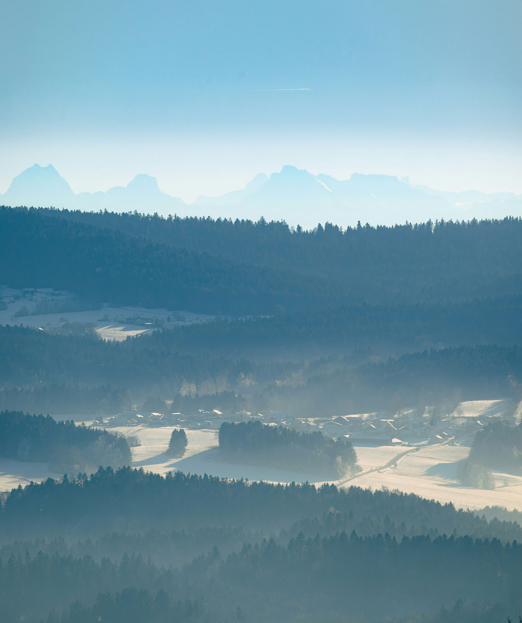 Verschneites Tal mit Dorf, Nadelwäldern und Alpenpanorama im Bayerischen Wald