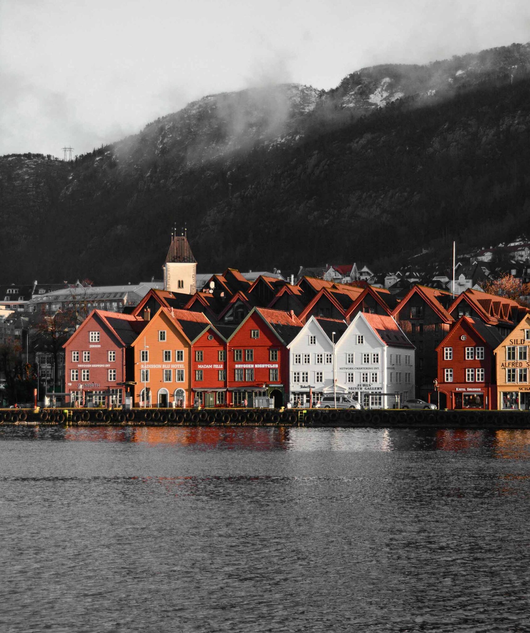 Bunte Holzhäuser des Hanseviertels Bryggen am Hafen von Bergen, dahinter bewaldete, wolkenverhangene Berge