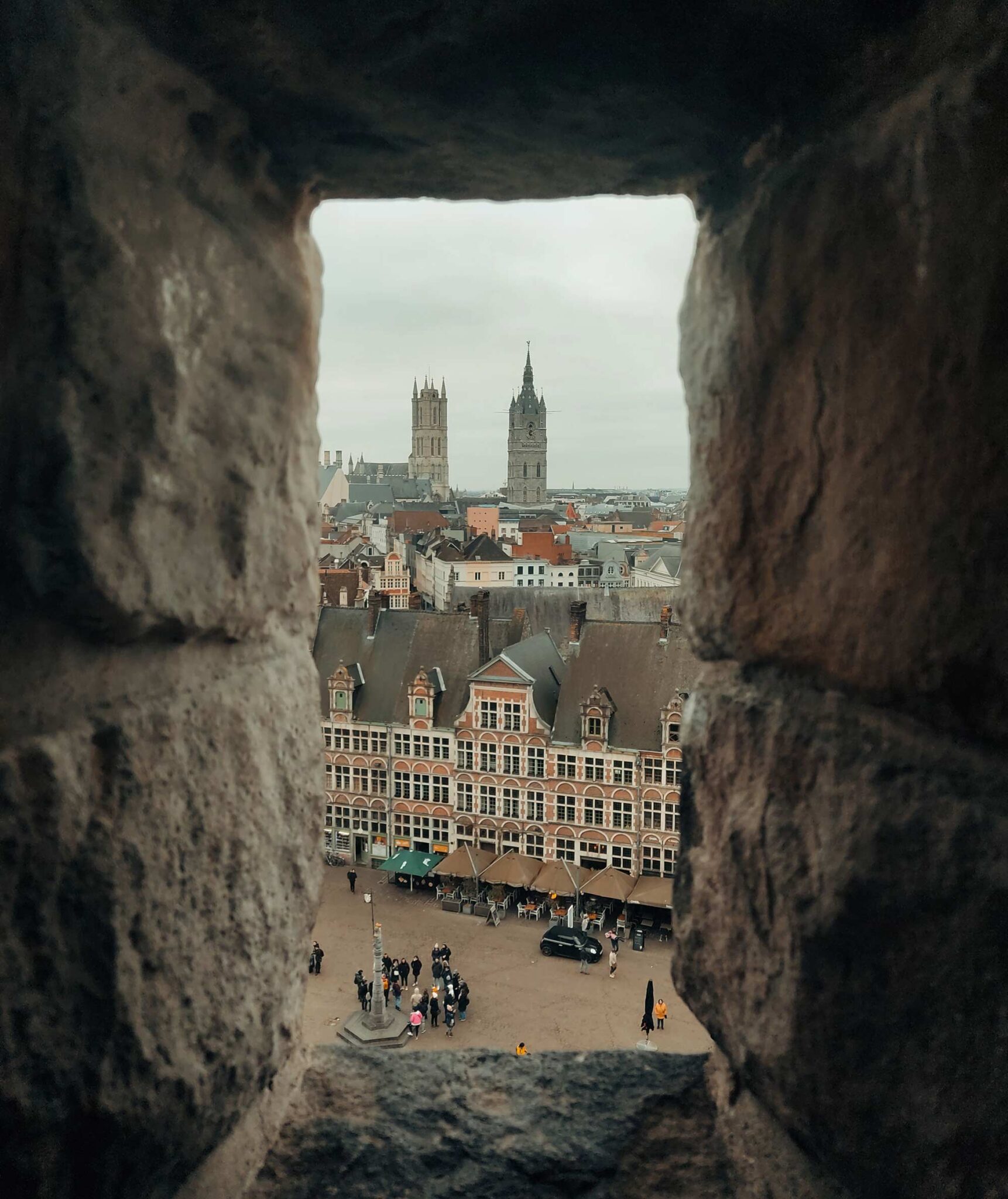 Blick durch eine Schießscharte der Burg Gravensteen auf historische Häuser und einen Platz in Gent, dahinter die Türme von Sankt-Bavo-Kathedrale und Belfried
